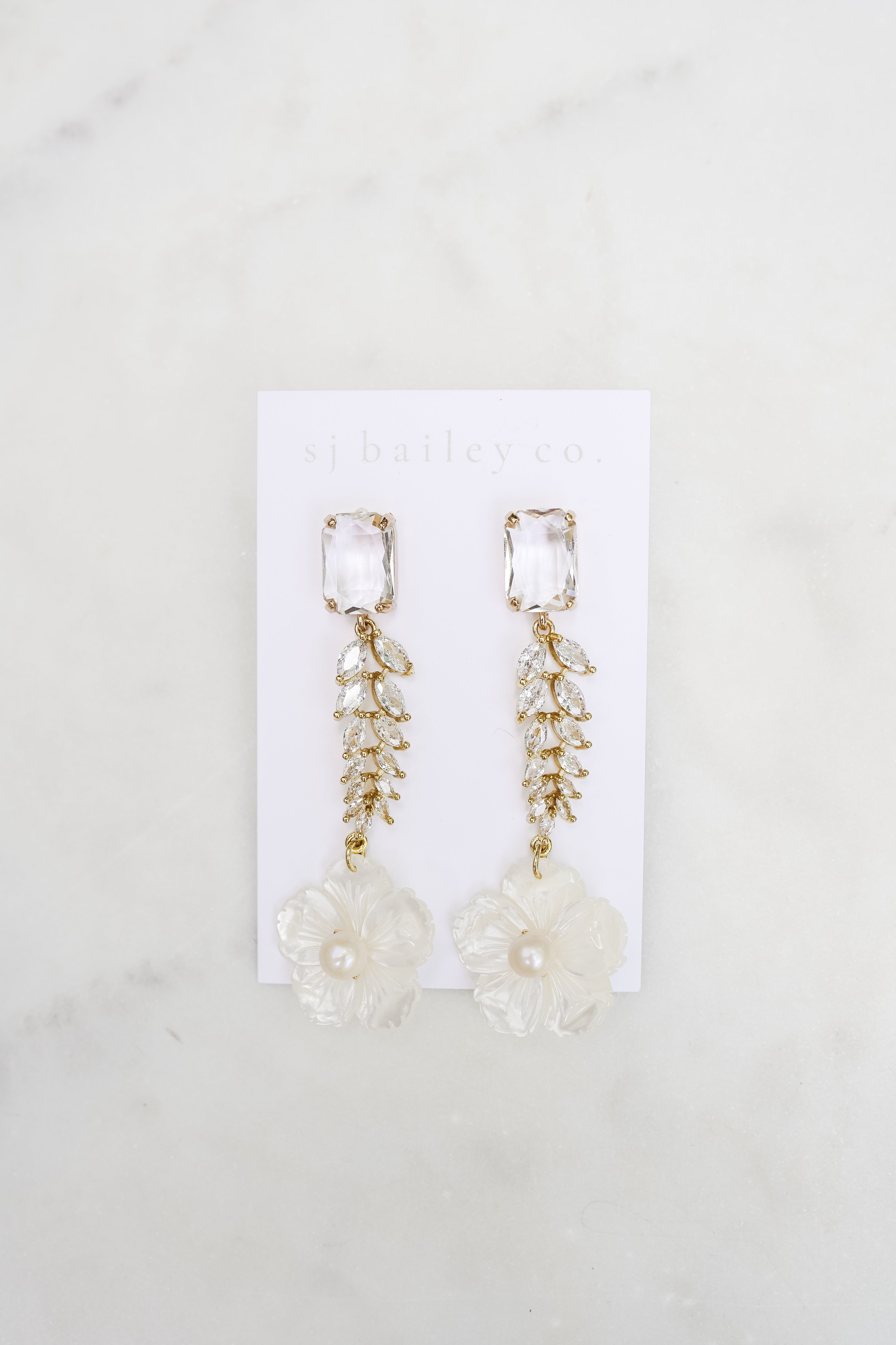Crystal earrings with a mother of pearl flower at the bottom. The earrings are on a white card on a marble background.
