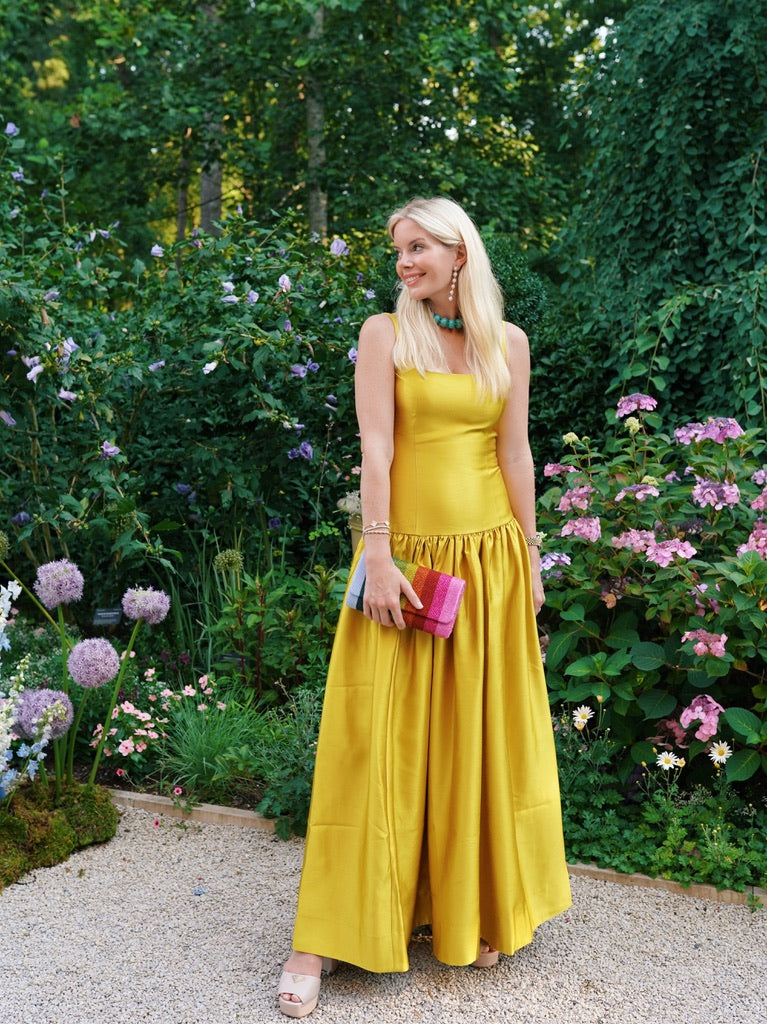A blonde woman wearing a yellow dress, blue necklace, rainbow clutch and sj bailey co floral earrings in front of a garden