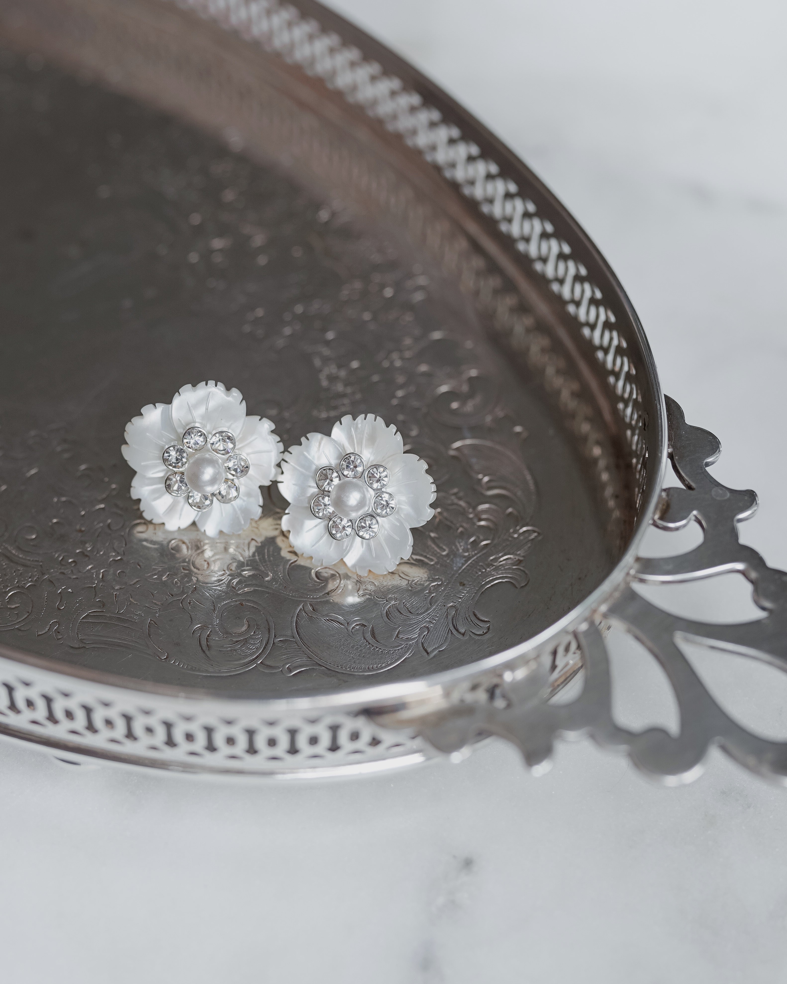 A pair of silver flower stud earrings with pearl-like centers, displayed on a decorative silver tray over a light marble surface.