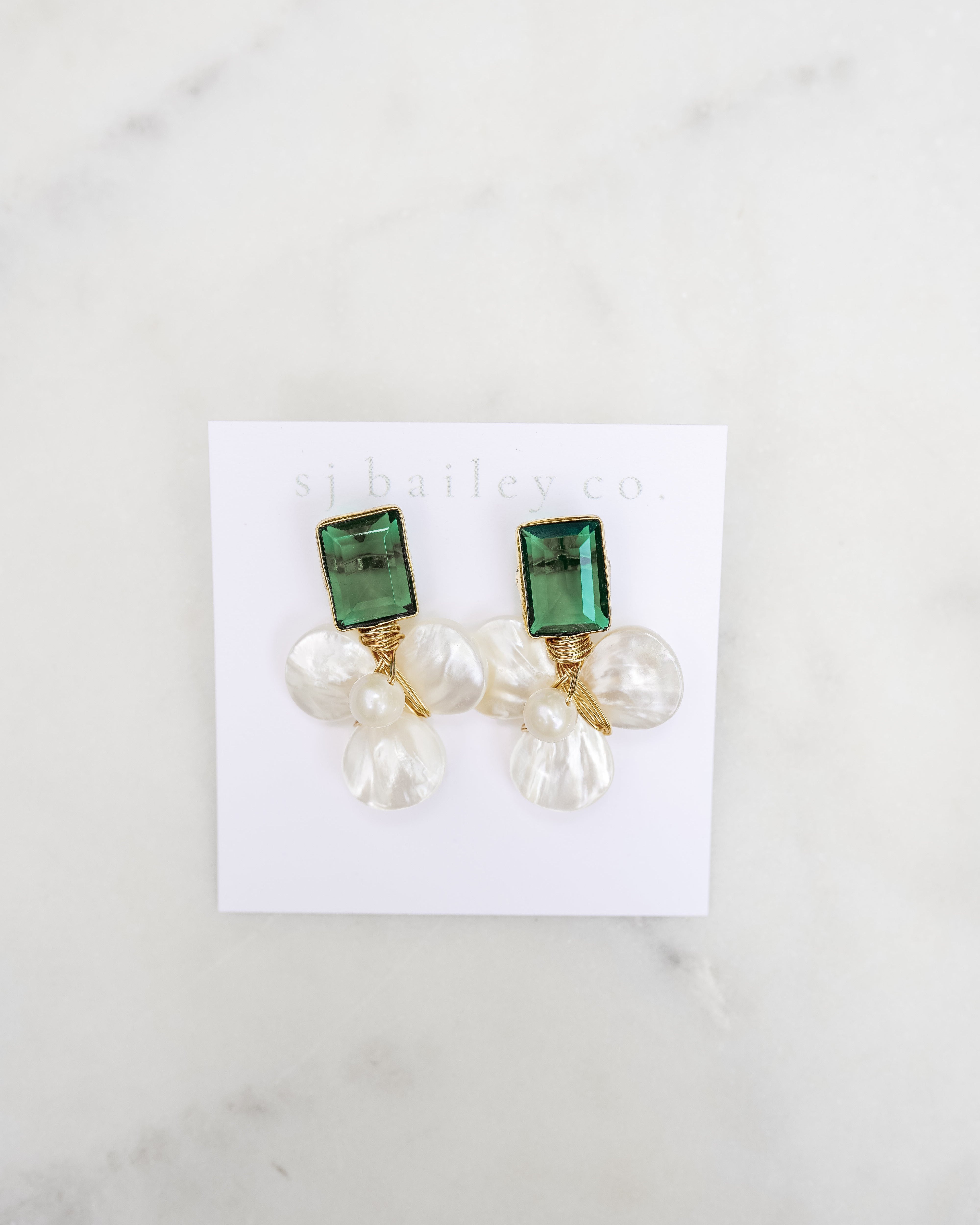 Dark emerald green and pearl earrings on a white card against a marble background.