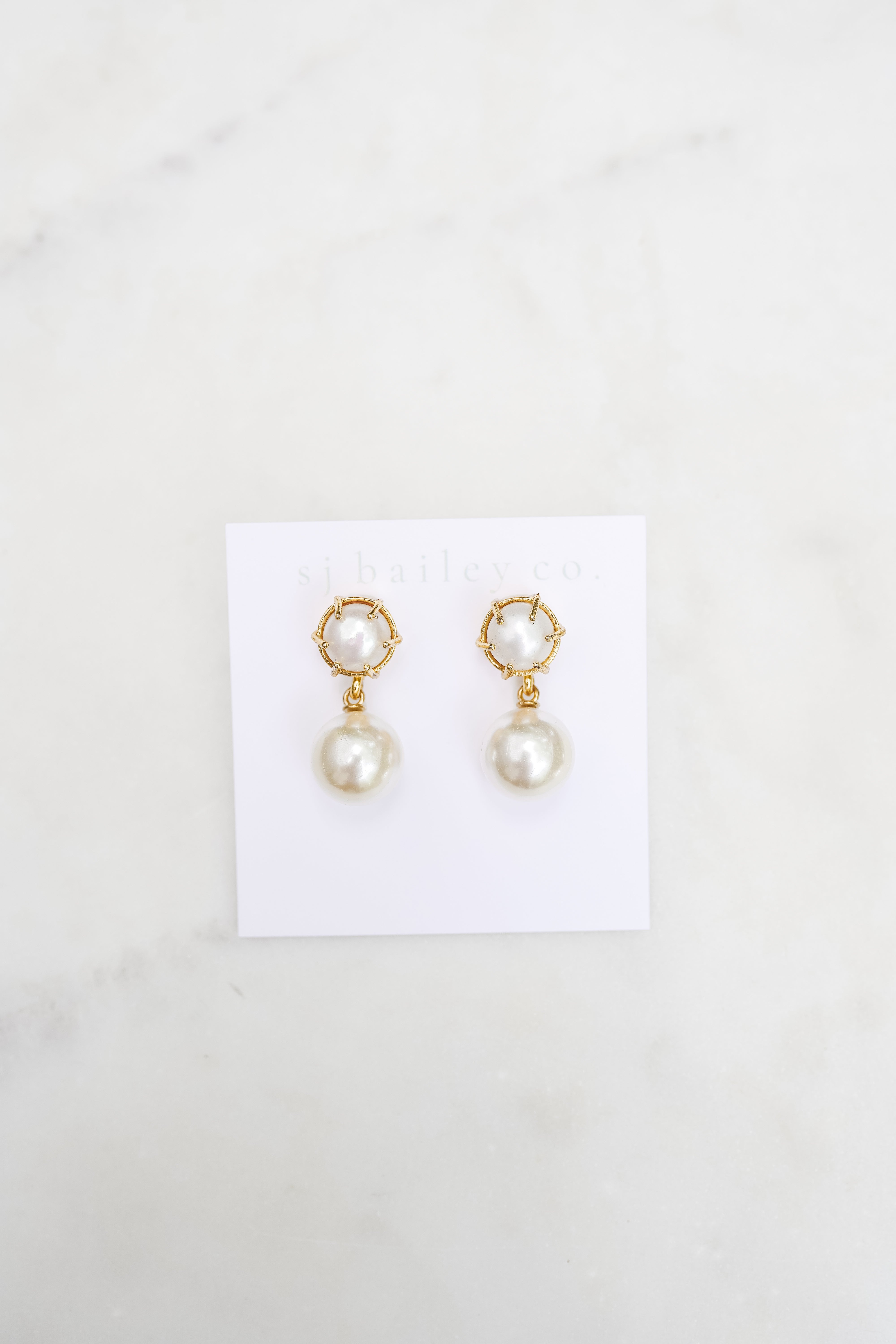 Pearl pendant earrings on a white card on a marble backdrop.