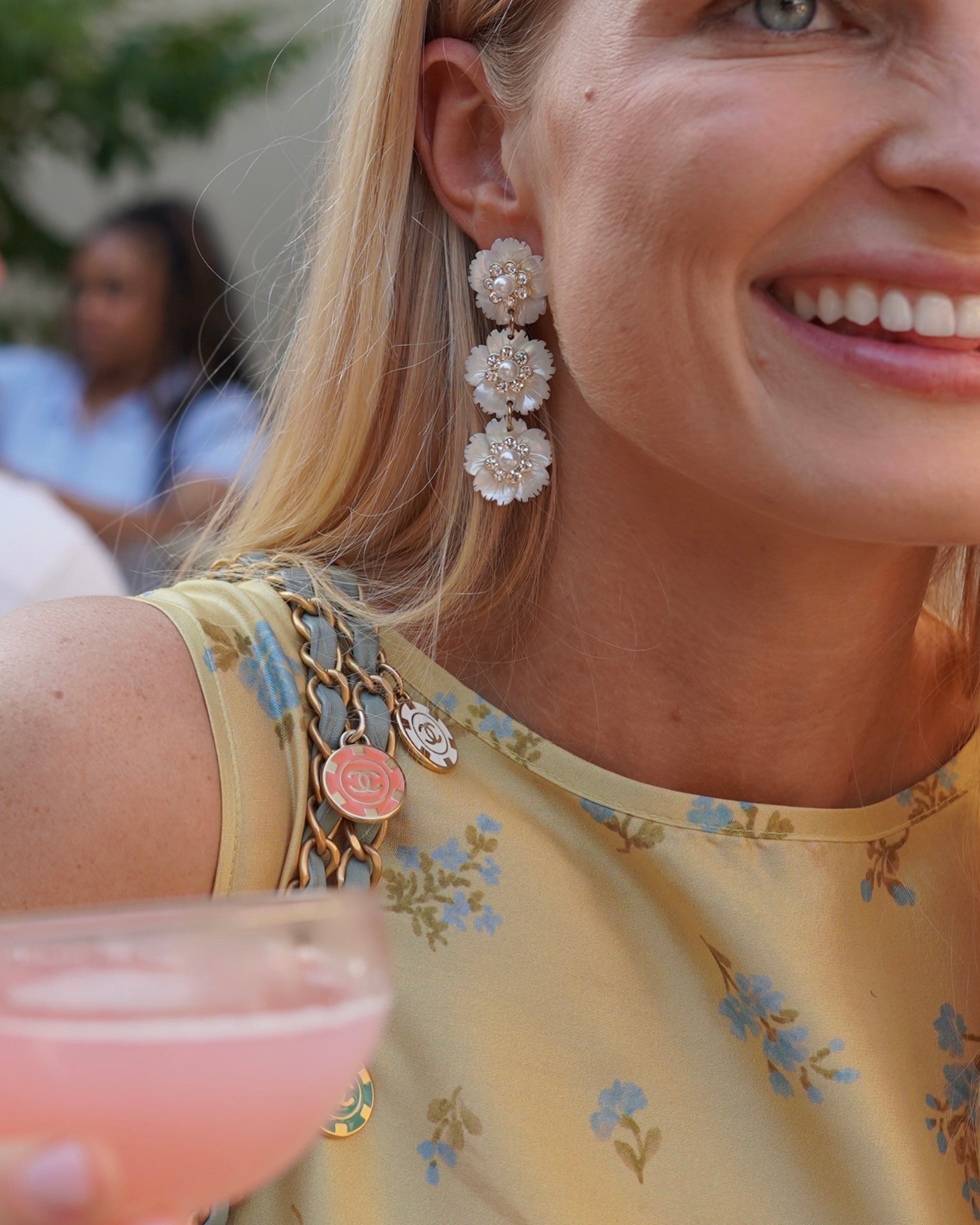 A close-up lifestyle shot of a woman wearing a pair of long, tiered dangle earrings made of white mother of pearl flowers and clear rhinestones, paired with a yellow floral dress.