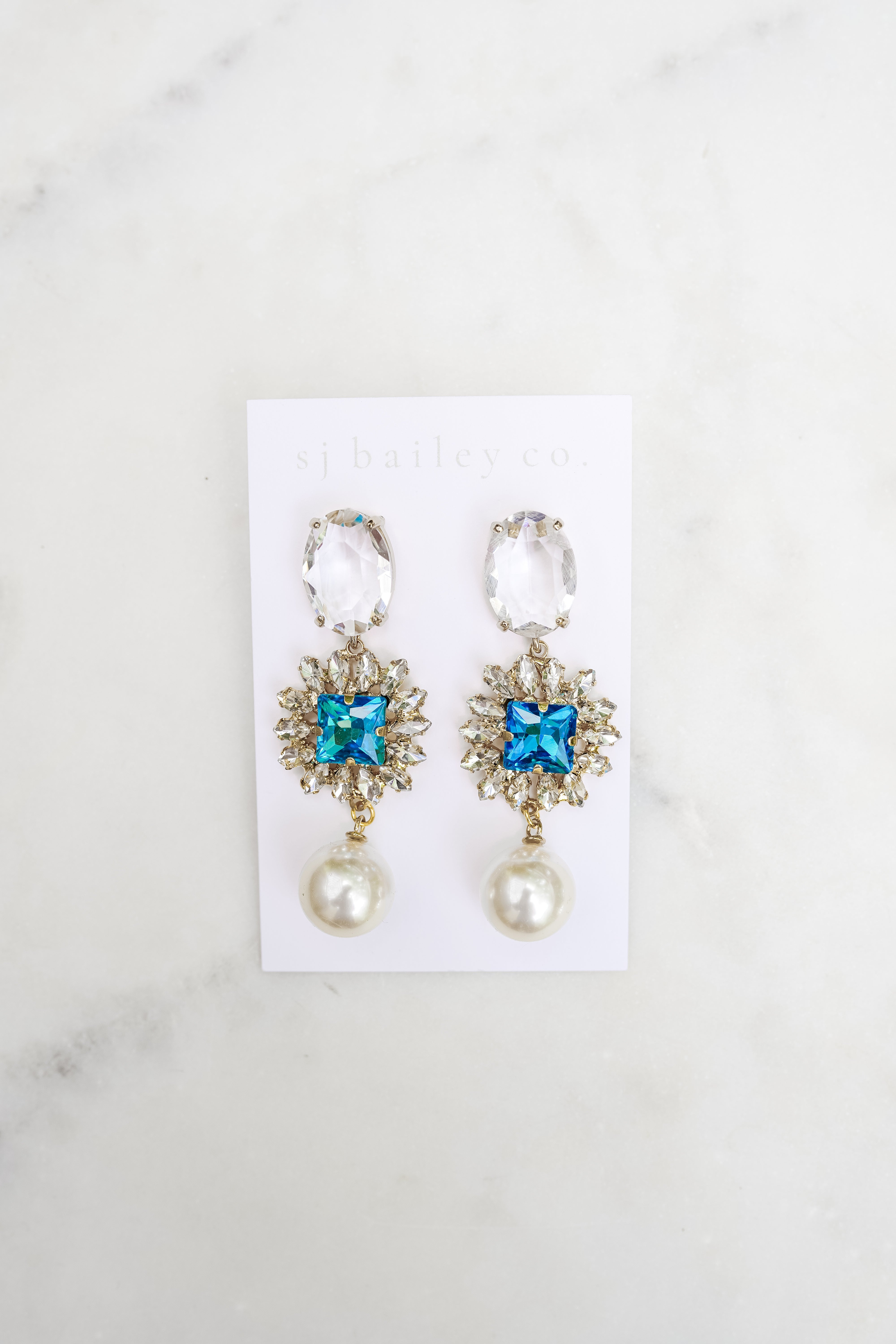 Pair of earrings with blue gemstones and pearl-like bottoms on a white card against a marble background.