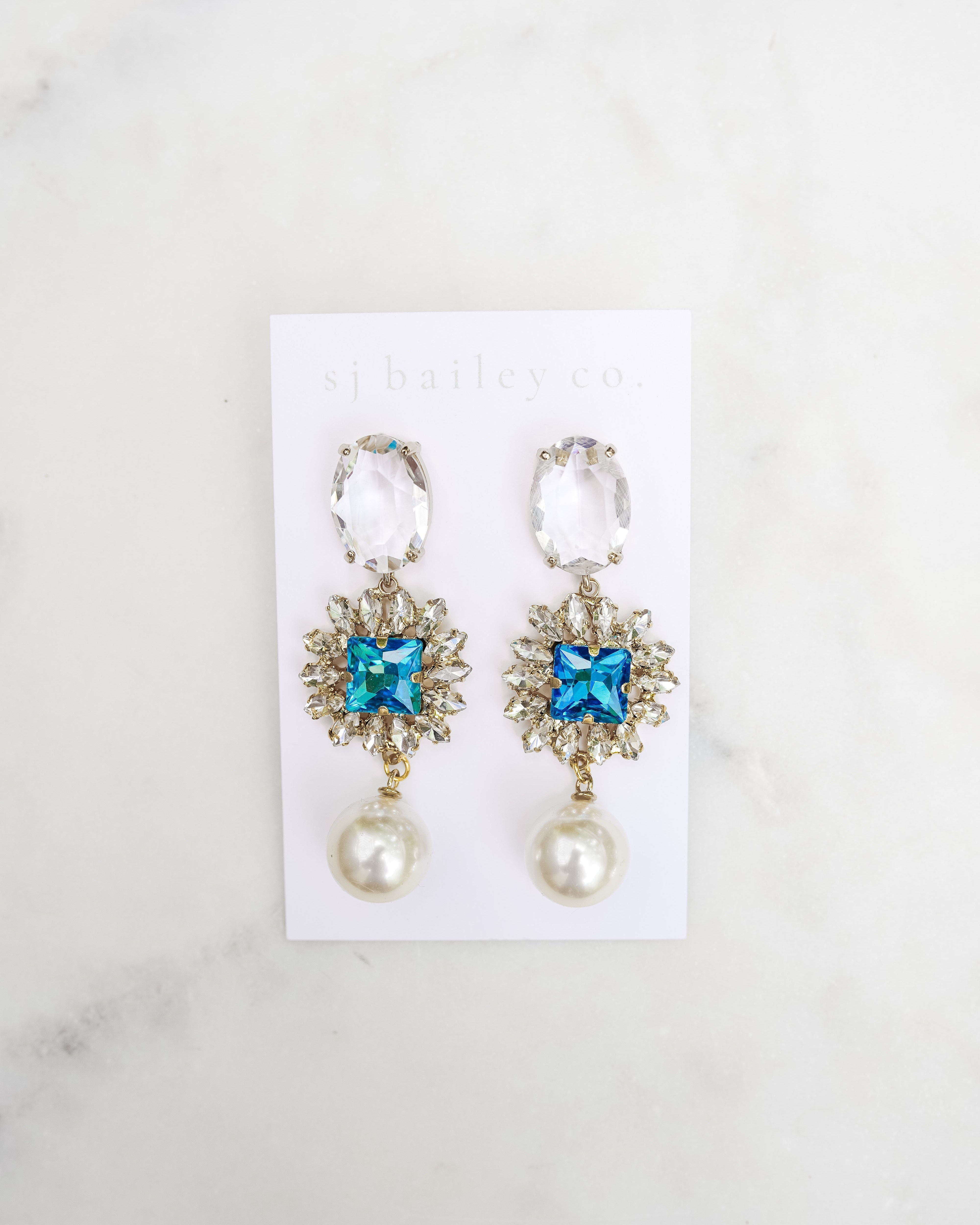 Pair of earrings with blue gemstones and pearl-like bottoms on a white card against a marble background.