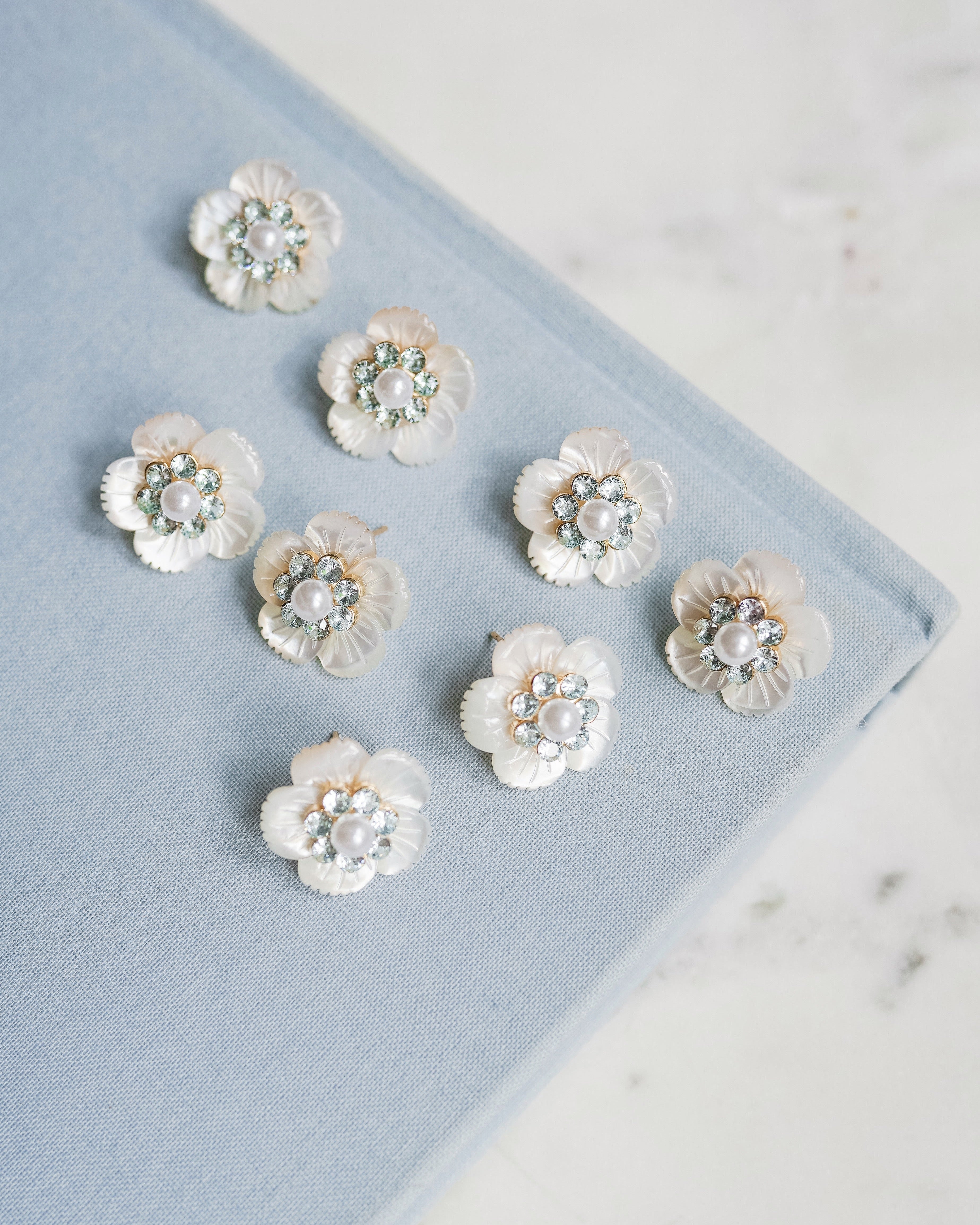 Eight white flower-shaped earrings with blue rhinestone centers, arranged on a light blue textured book cover against a white marble background.