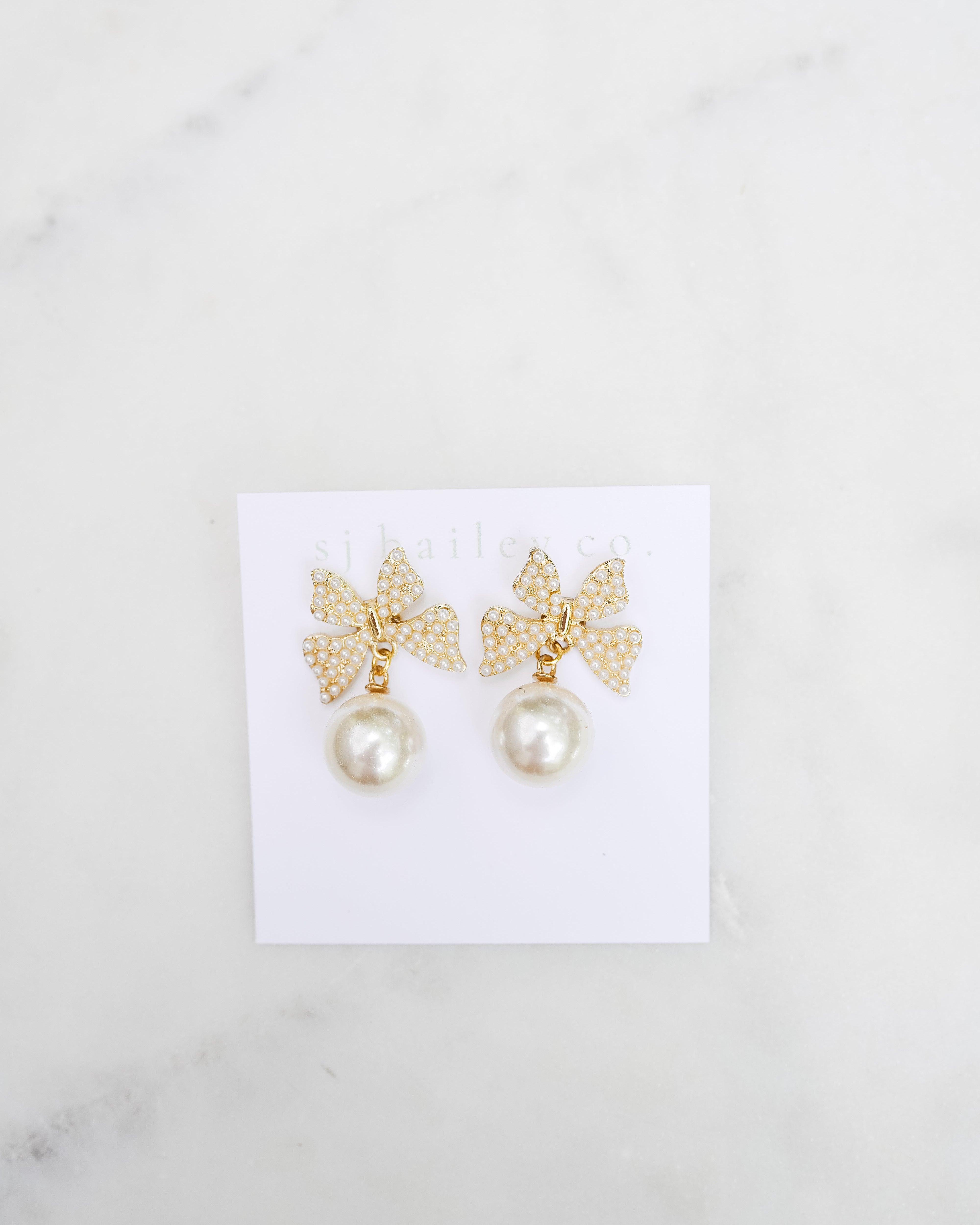 Golden and Pearl bow earrings with a pearl pendant on a white card. They are featured on a marble background.