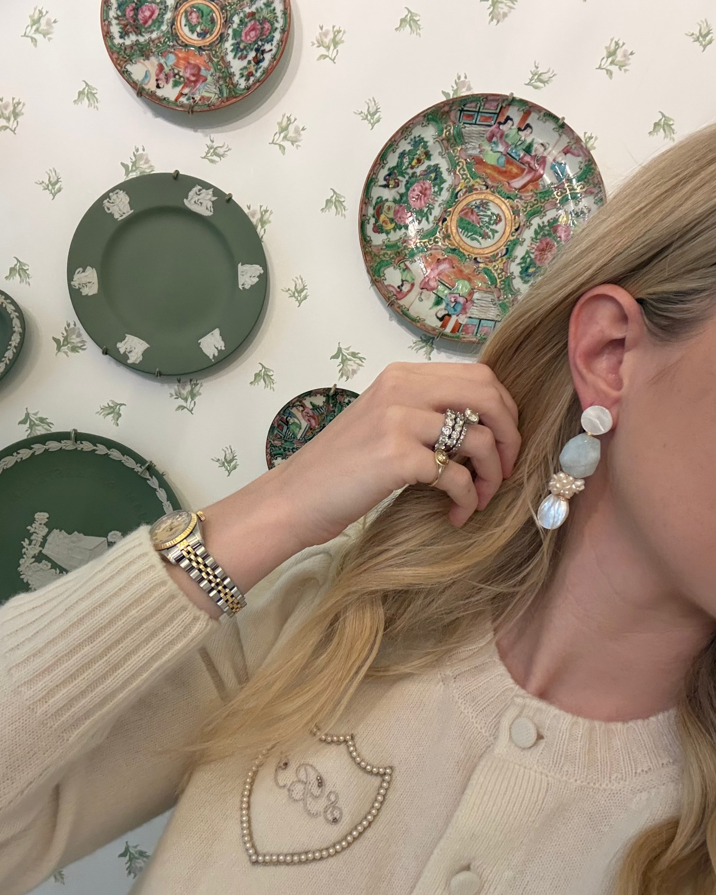 a blonde woman in a cream sweater wearing aquamarine and pearl statement earrings. she is in front of a floral wallpapered wall with green wedgwood and rose medallion plates on it