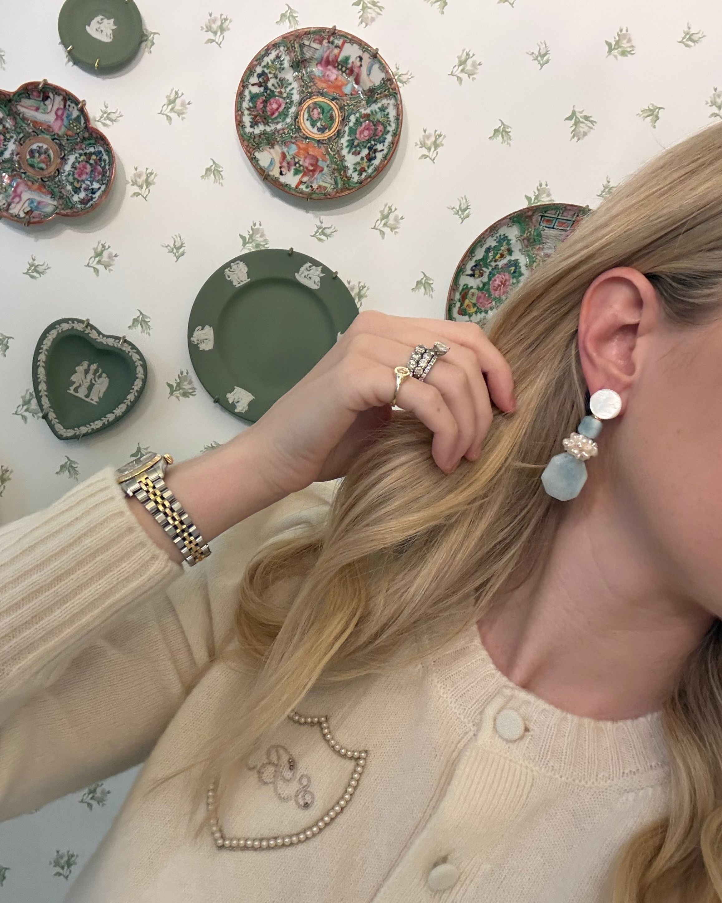 a blonde woman in a cream sweater wearing aquamarine and pearl statement earrings. she is in front of a floral wallpapered wall with green wedgwood and rose medallion plates on it