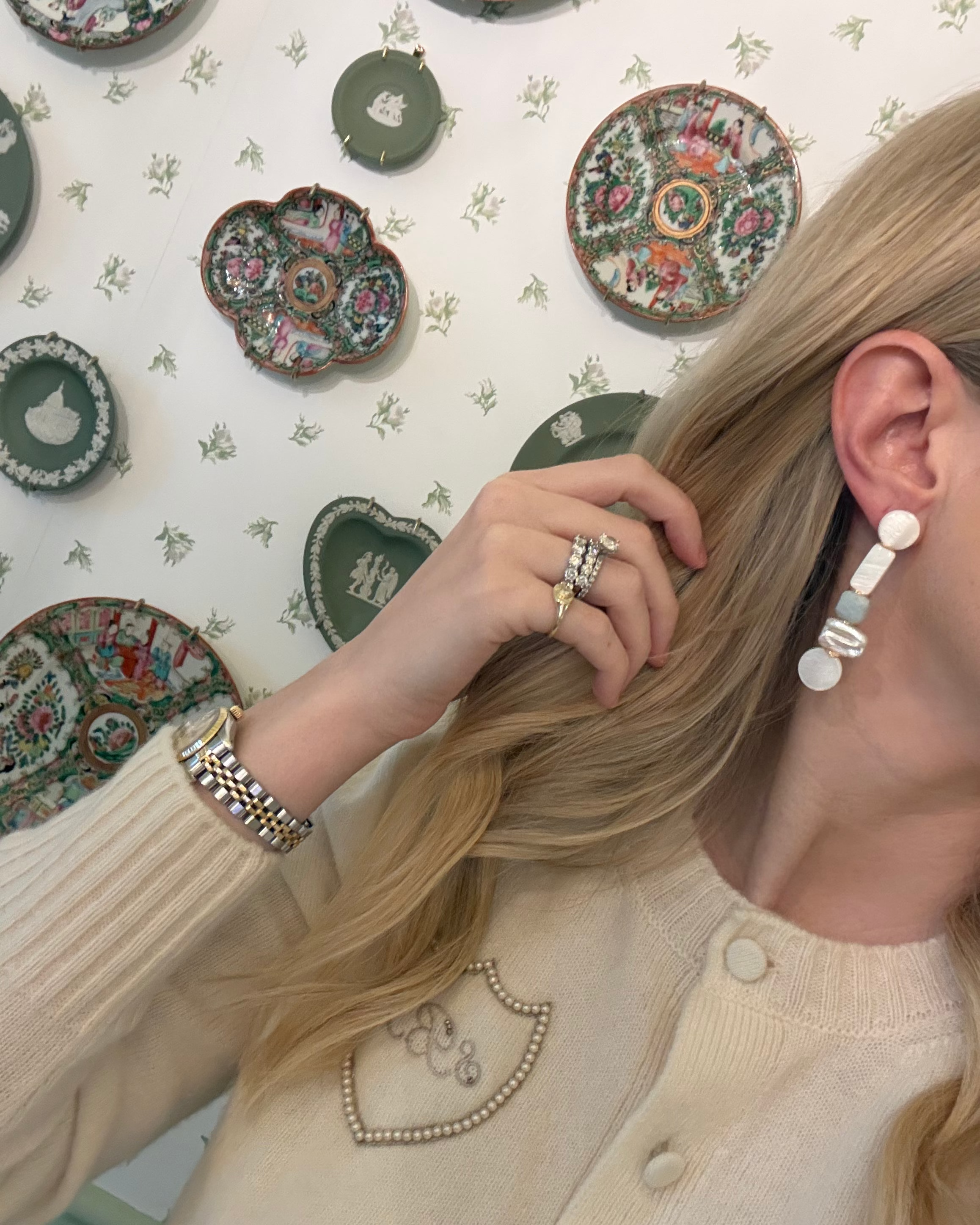 a blonde woman in a cream sweater wearing aquamarine and pearl statement earrings. she is in front of a floral wallpapered wall with green wedgwood and rose medallion plates on it