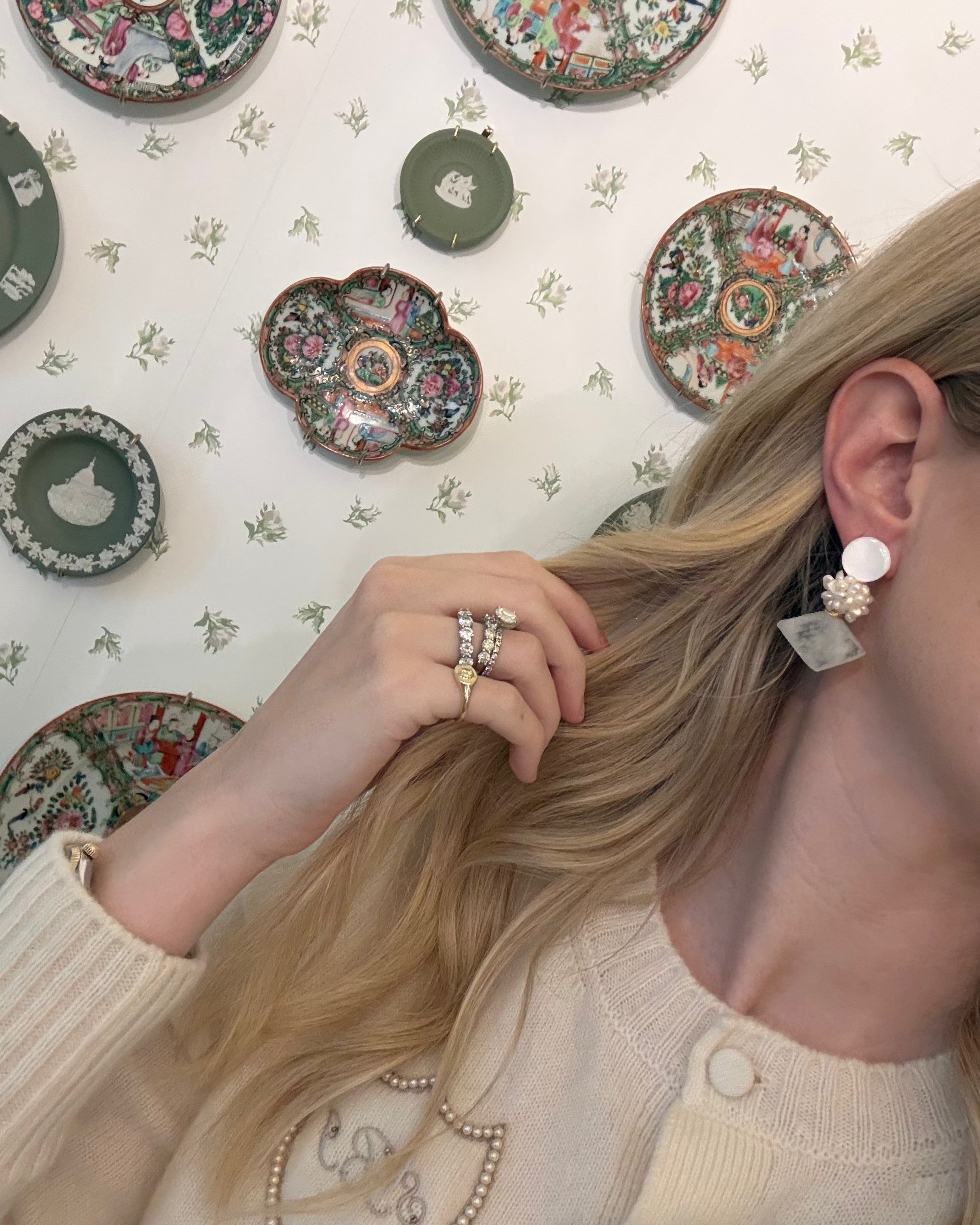 a blonde woman in a cream sweater wearing aquamarine and pearl statement earrings. she is in front of a floral wallpapered wall with green wedgwood and rose medallion plates on it