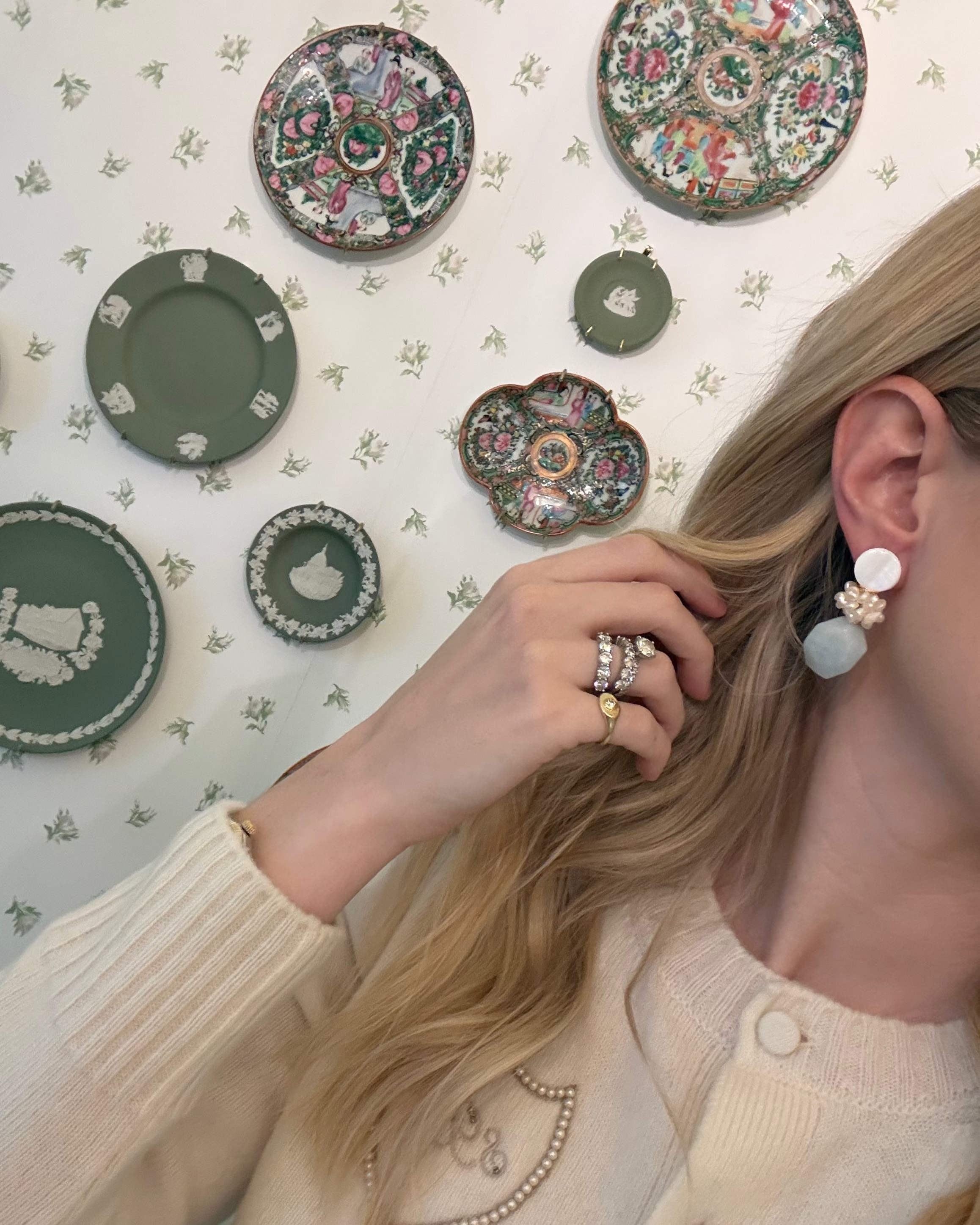 a blonde woman in a cream sweater wearing aquamarine and pearl statement earrings. she is in front of a floral wallpapered wall with green wedgwood and rose medallion plates on it