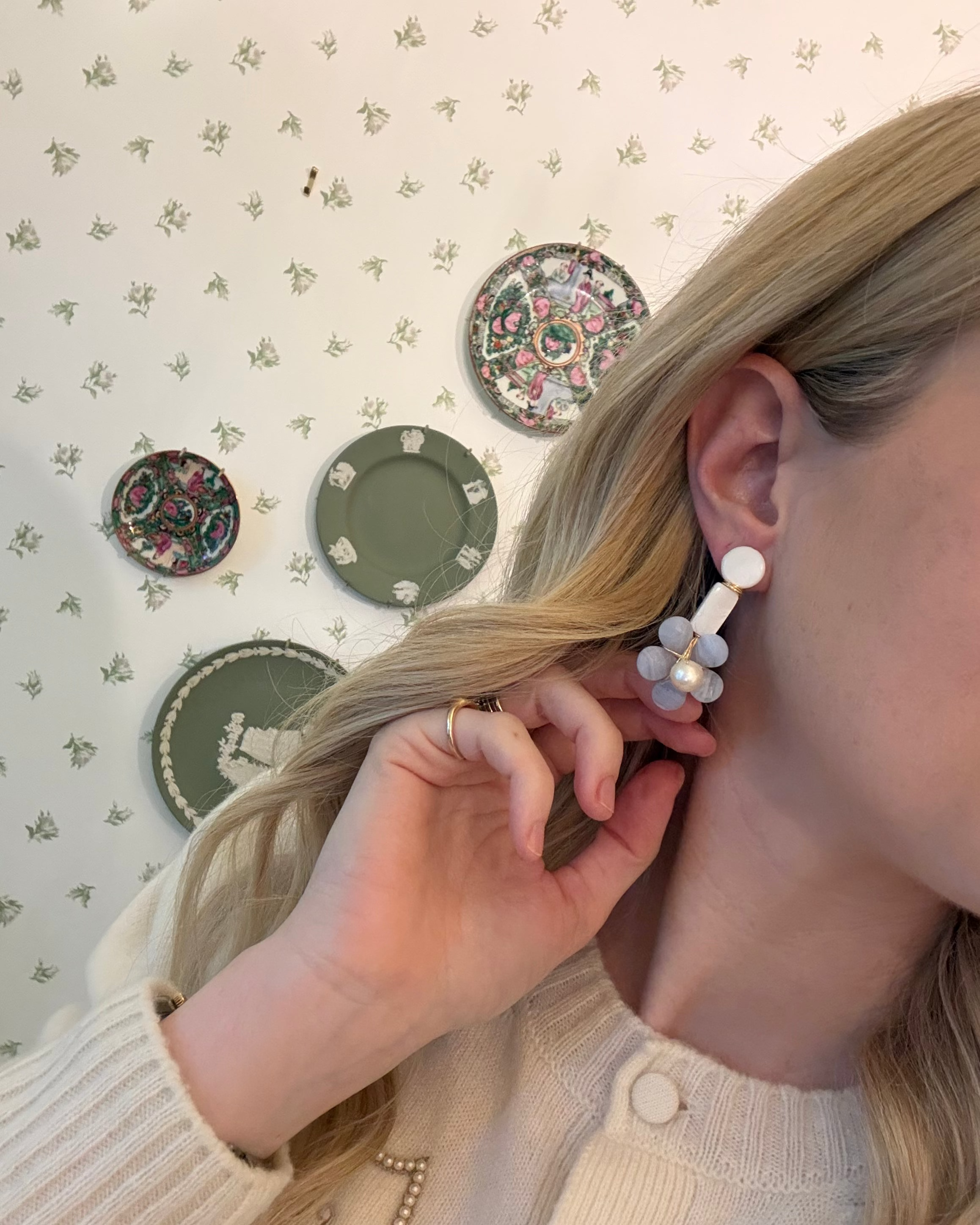a blonde woman in a cream sweater wearing aquamarine and pearl statement earrings. she is in front of a floral wallpapered wall with green wedgwood and rose medallion plates on it