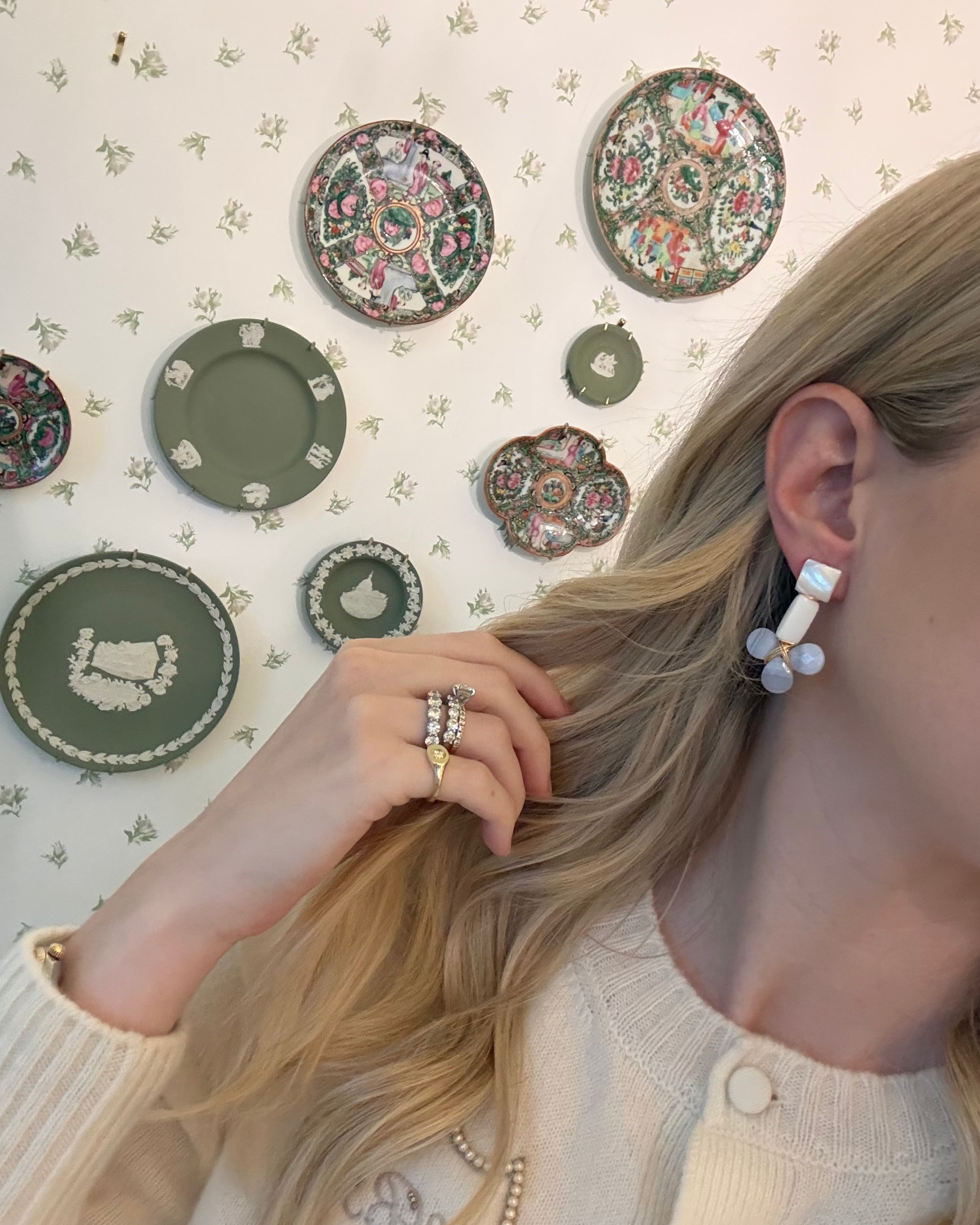 a blonde woman in a cream sweater wearing aquamarine and pearl statement earrings. she is in front of a floral wallpapered wall with green wedgwood and rose medallion plates on it