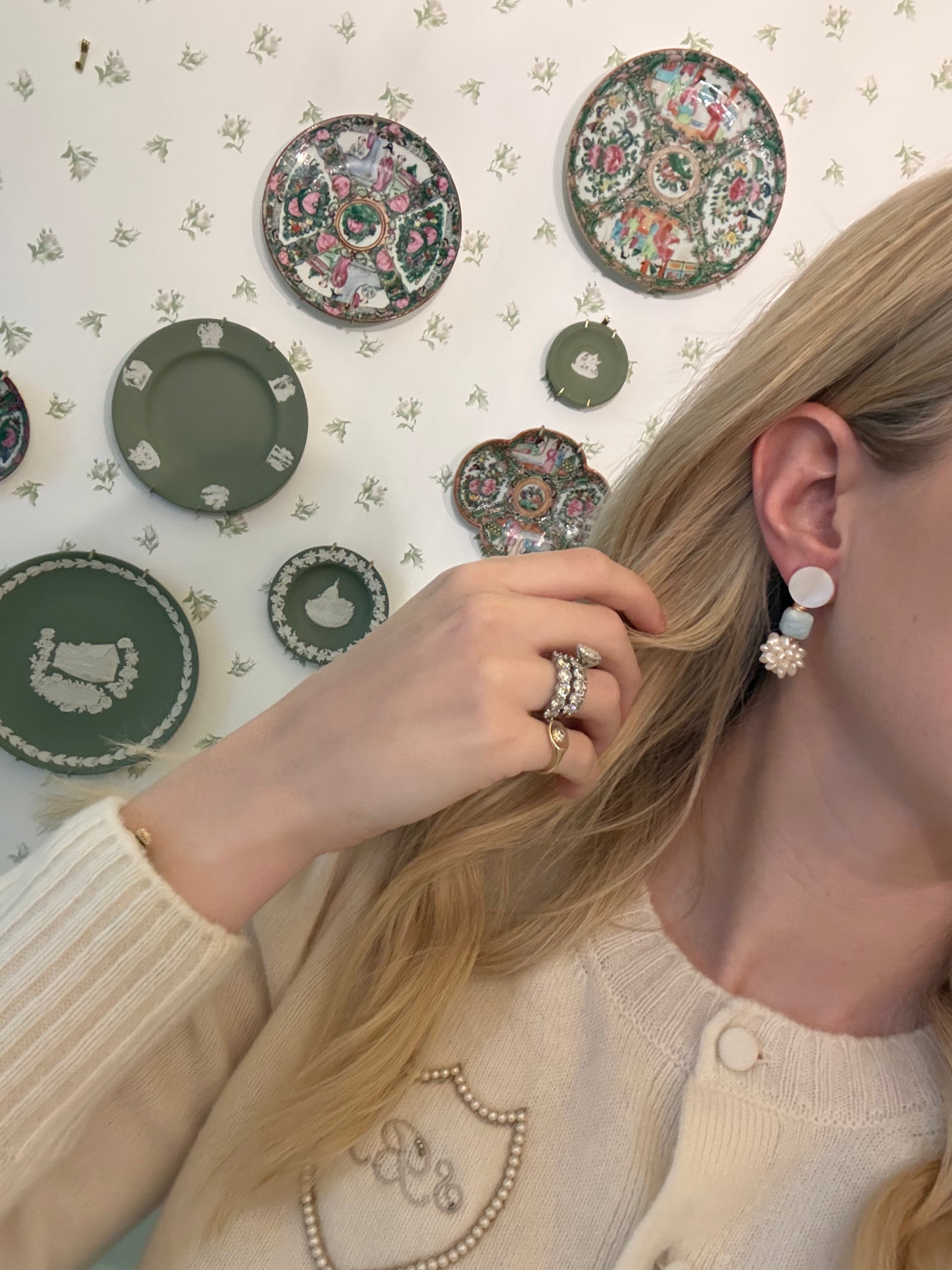 a blonde woman in a cream sweater wearing aquamarine and pearl statement earrings. she is in front of a floral wallpapered wall with green wedgwood and rose medallion plates on it