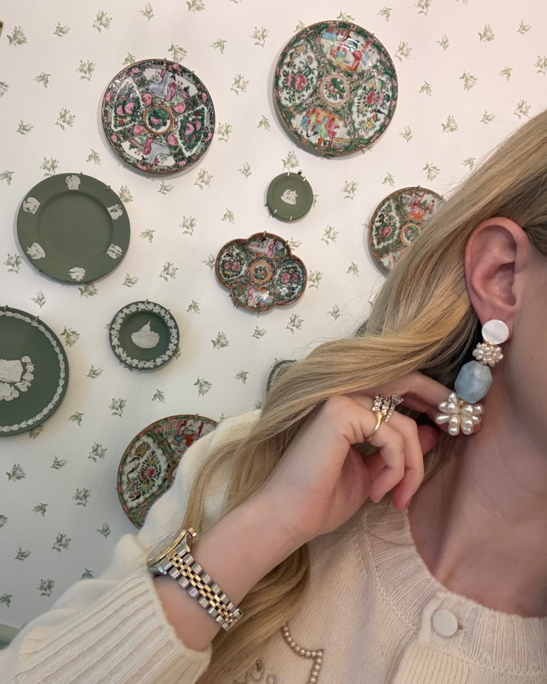 a blonde woman in a cream sweater wearing aquamarine and pearl statement earrings. she is in front of a floral wallpapered wall with green wedgwood and rose medallion plates on it