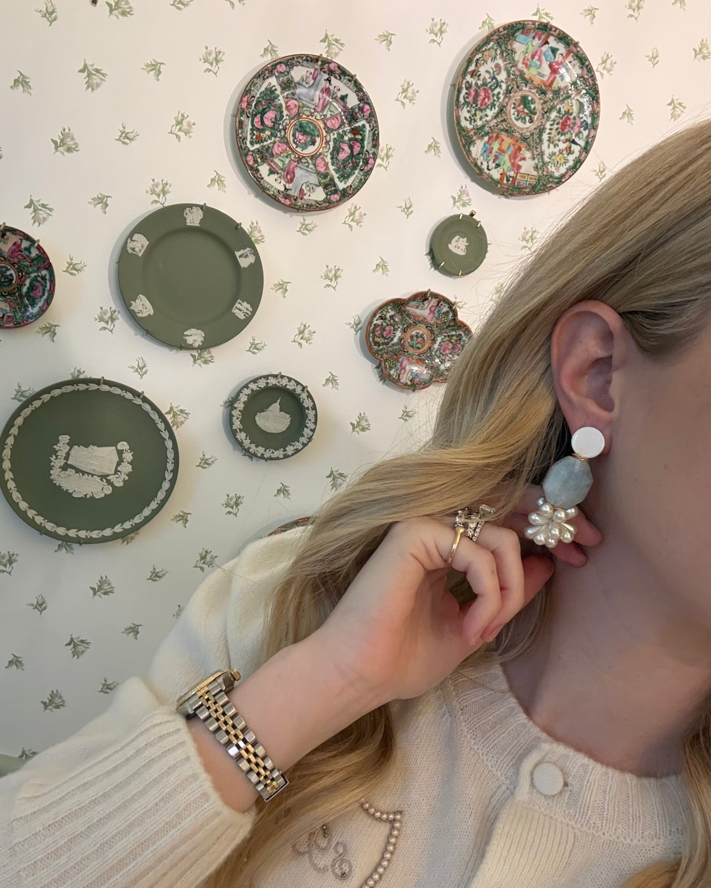 a blonde woman in a cream sweater wearing aquamarine and pearl statement earrings. she is in front of a floral wallpapered wall with green wedgwood and rose medallion plates on it