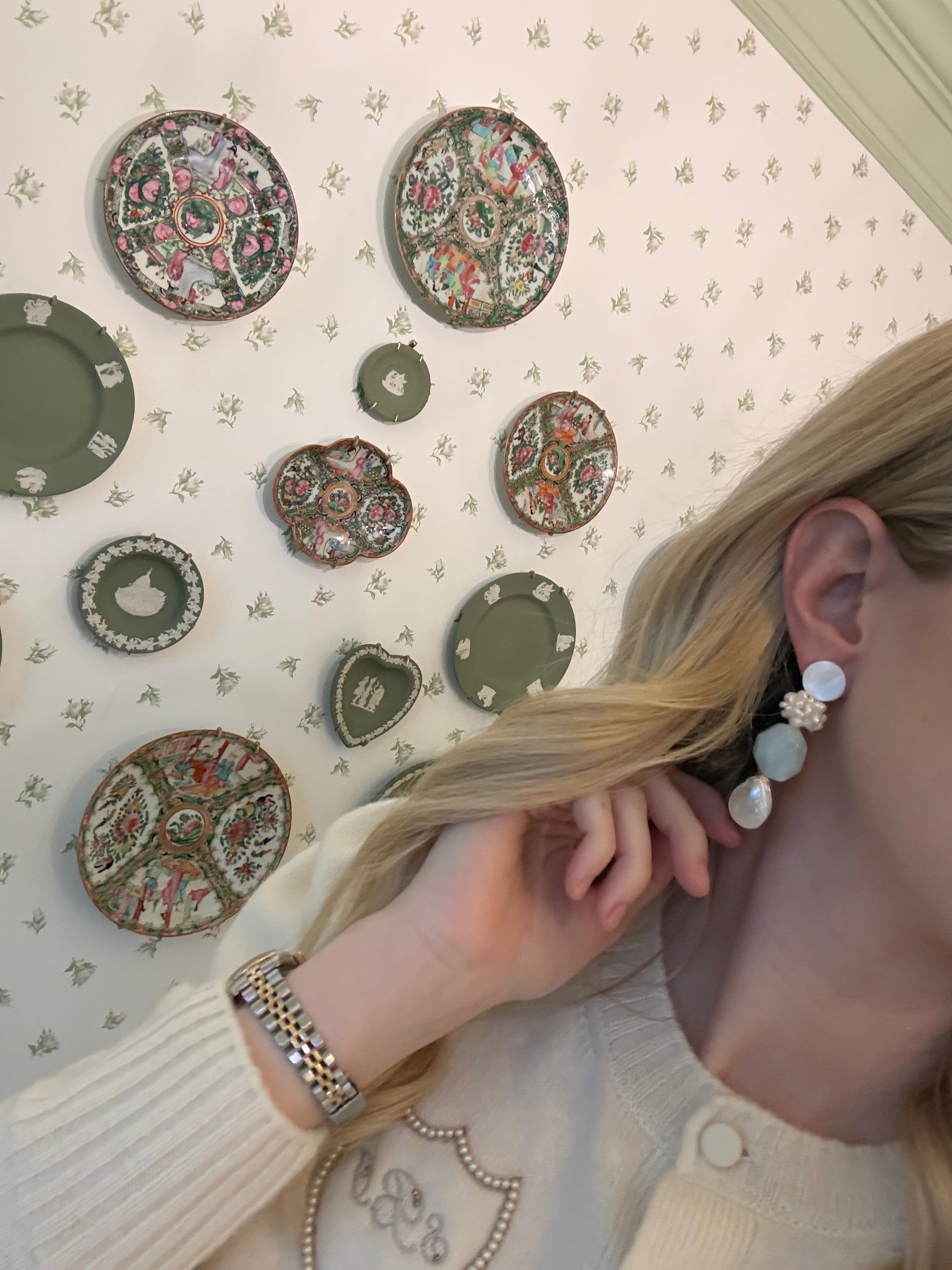 a blonde woman in a cream sweater wearing aquamarine and pearl statement earrings. she is in front of a floral wallpapered wall with green wedgwood and rose medallion plates on it