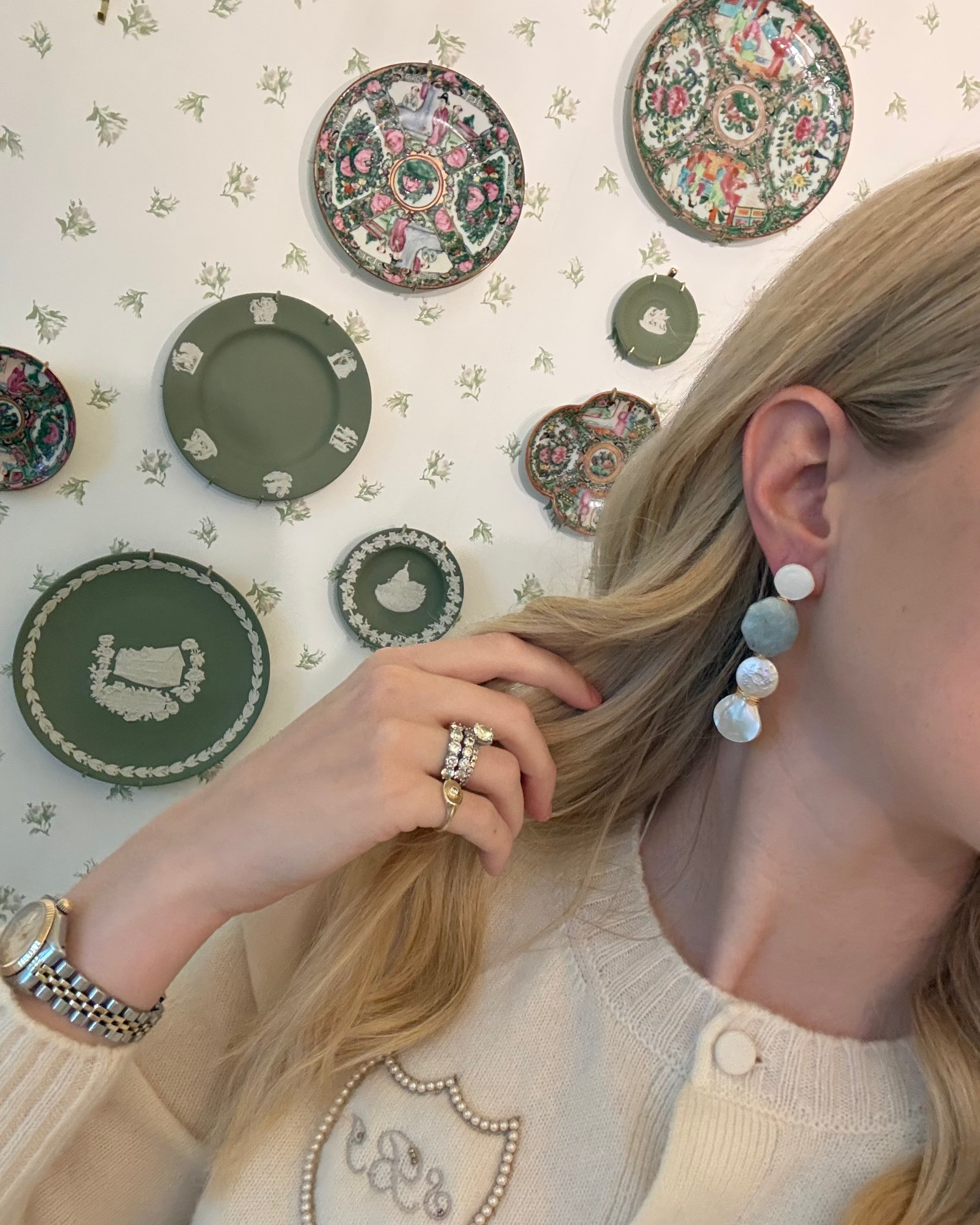 a blonde woman in a cream sweater wearing aquamarine and pearl statement earrings. she is in front of a floral wallpapered wall with green wedgwood and rose medallion plates on it