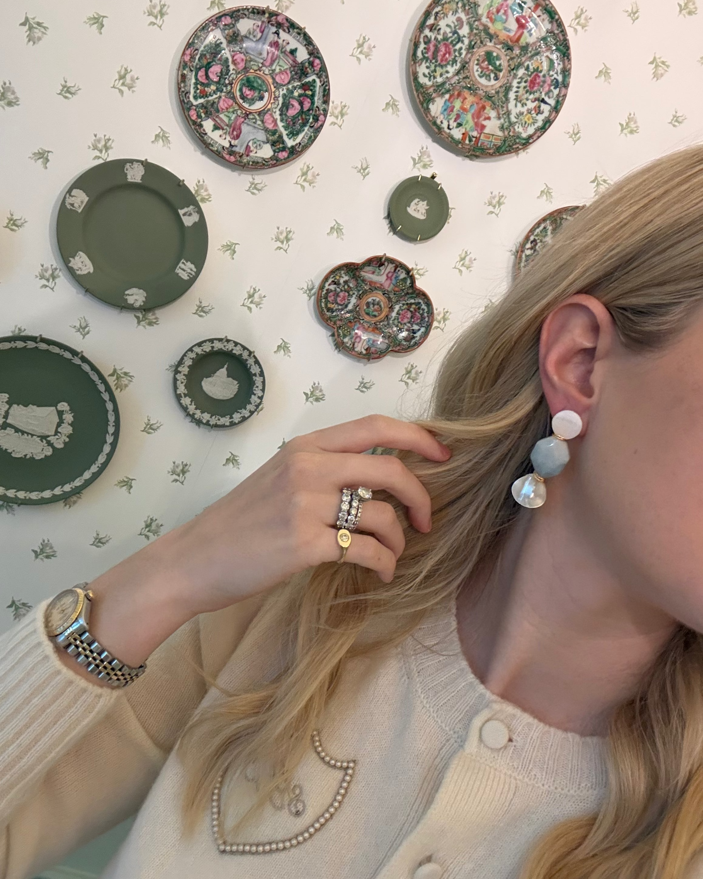 a blonde woman in a cream sweater wearing aquamarine and pearl statement earrings. she is in front of a floral wallpapered wall with green wedgwood and rose medallion plates on it