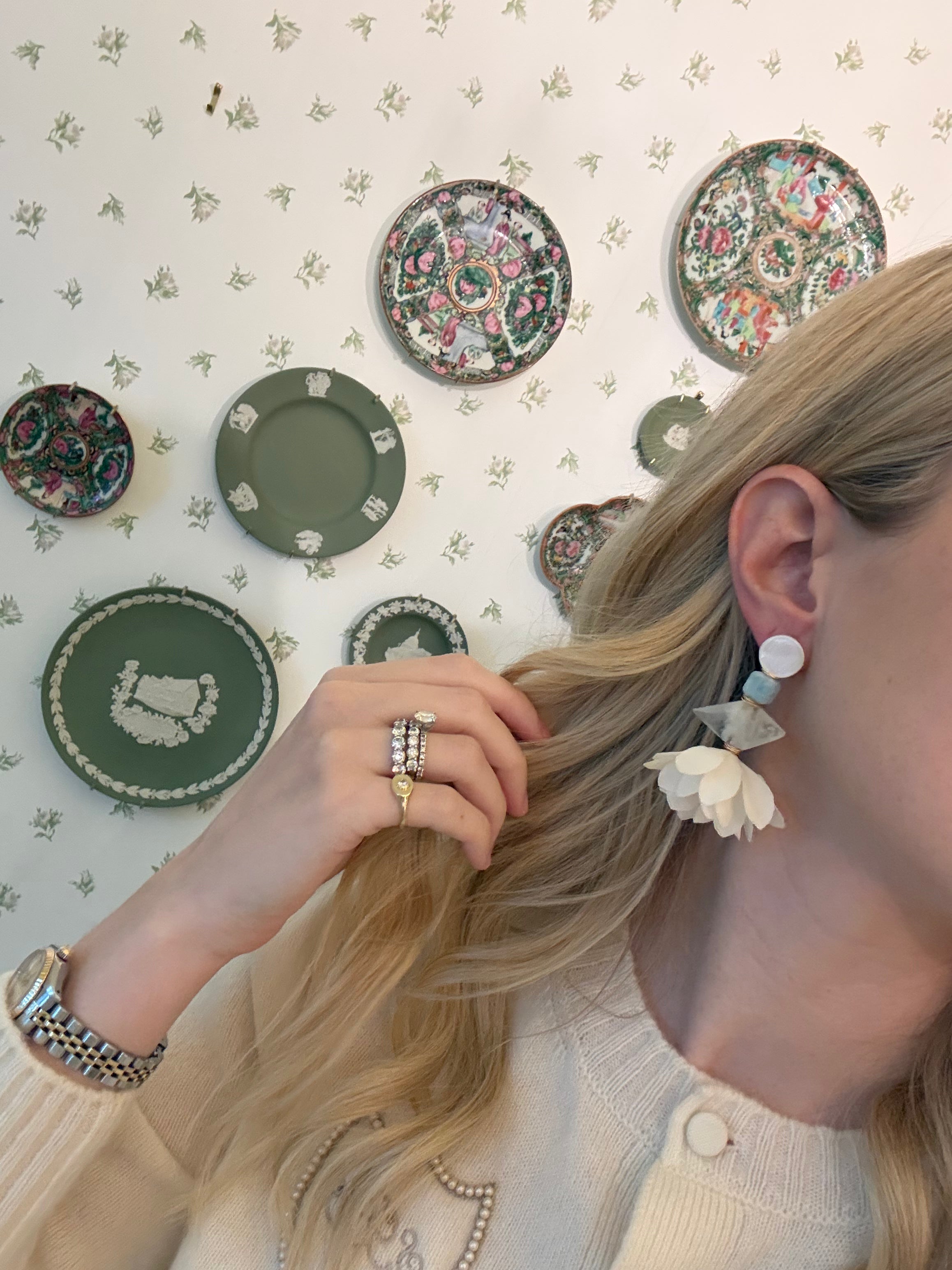 a blonde woman in a cream sweater wearing aquamarine and pearl statement earrings. she is in front of a floral wallpapered wall with green wedgwood and rose medallion plates on it