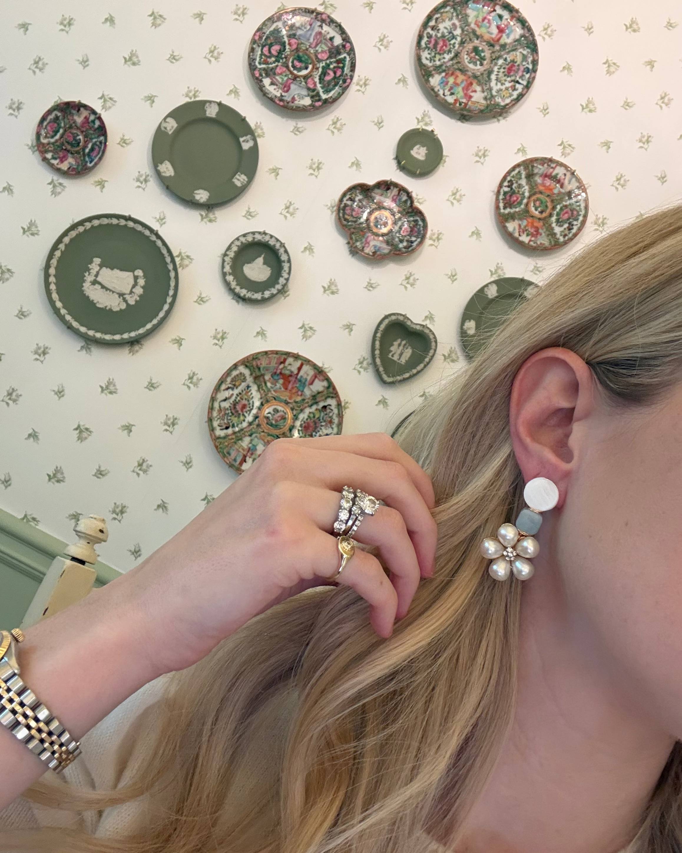 a blonde woman in a cream sweater wearing aquamarine and pearl statement earrings. she is in front of a floral wallpapered wall with green wedgwood and rose medallion plates on it