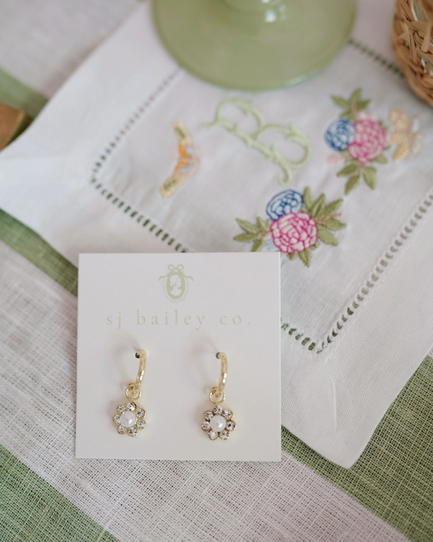 Gold hoop earrings with an embellished crystal and pearl flower on a green and white stripe tablecloth and embroidered floral napkin