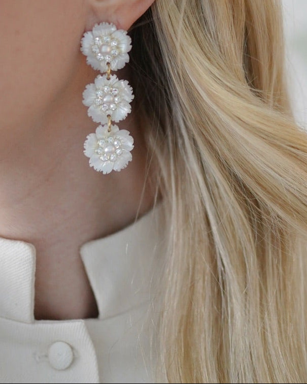 A close-up lifestyle shot of a blonde woman wearing a long, three-tier dangle earring made of white mother of pearl flowers, paired with a cream-colored high-neck top. 