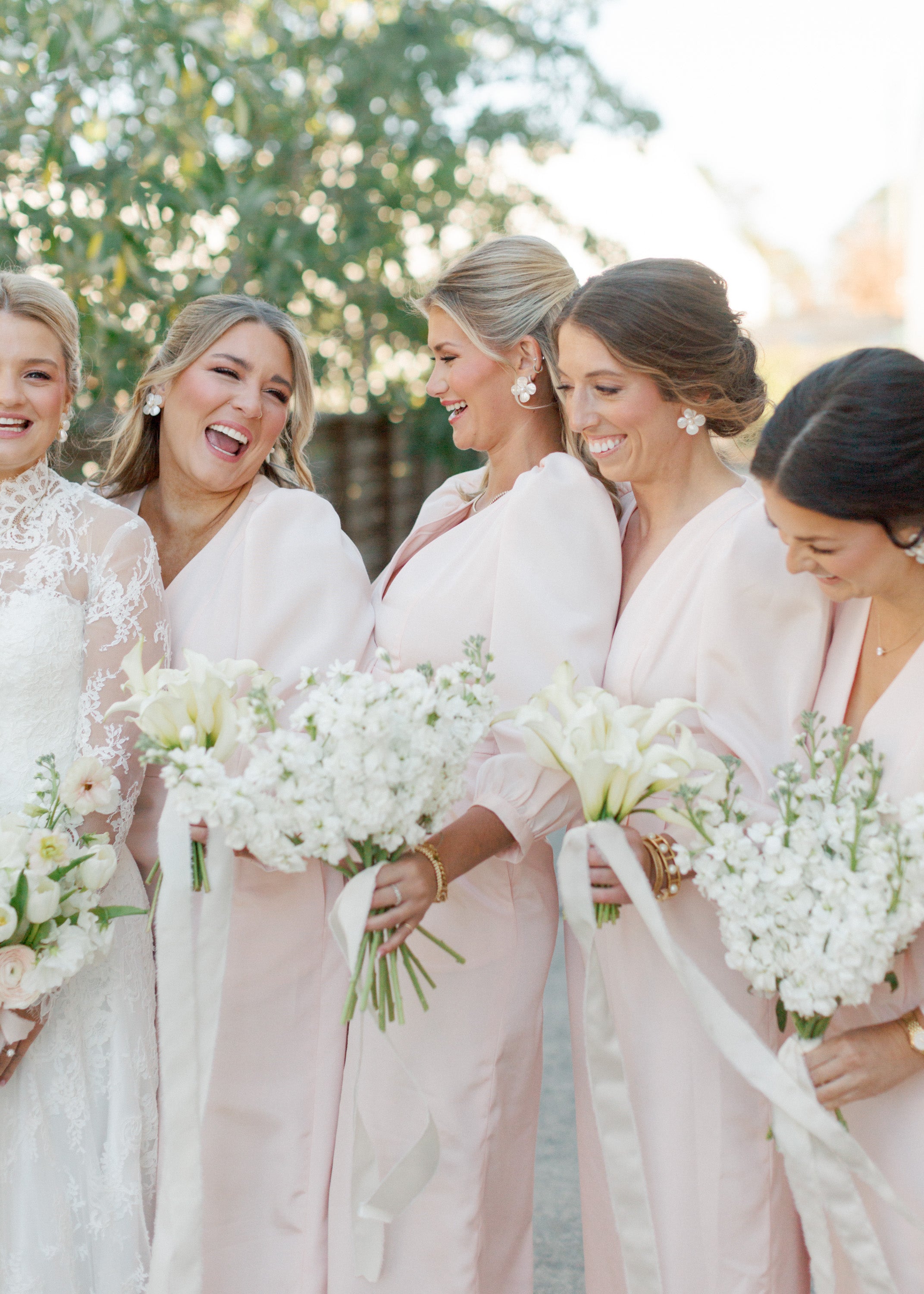 Group of women in light pink dresses holding white bouquets outdoors.