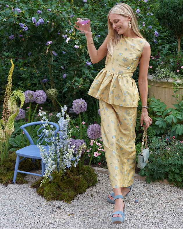 A woman stands in a lush garden wearing a yellow floral peplum top and matching skirt, holding a small blue handbag.