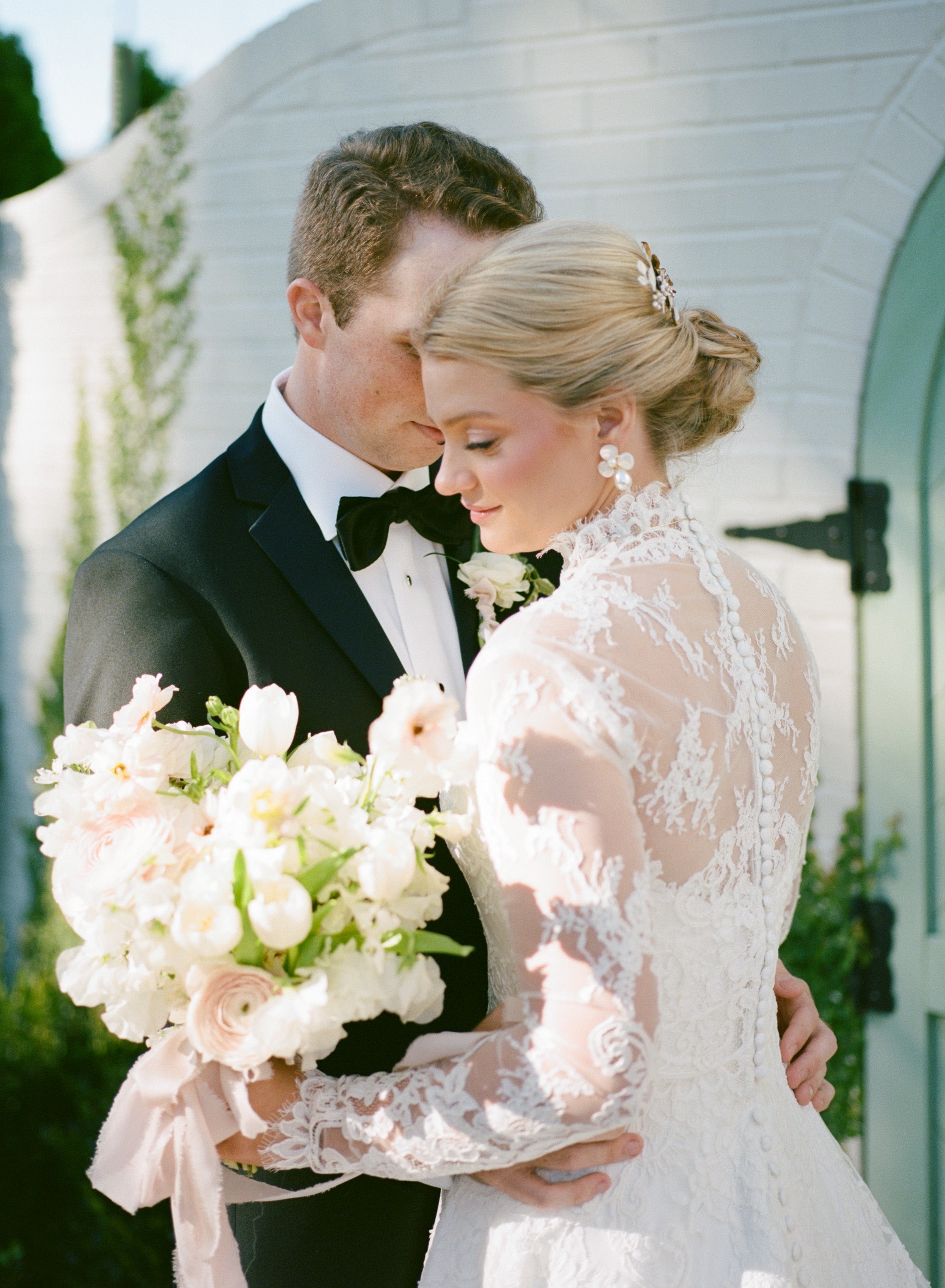 Man in tuxedo and woman in lace wedding dress holding a bouquet, standing in front of a white arch.