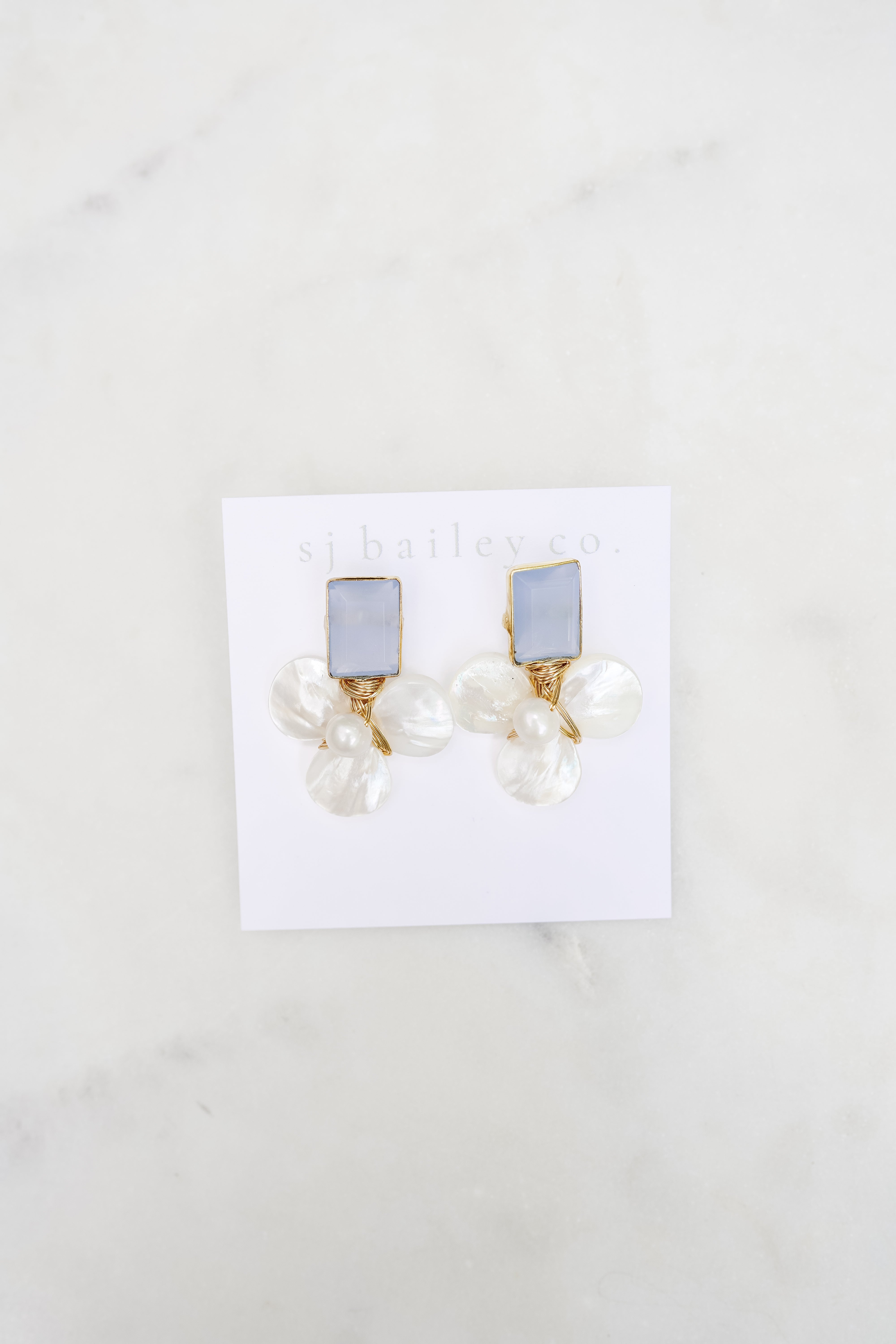 Dusty blue and pearl earrings against a white card on a marble background. 