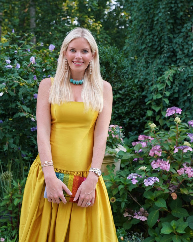 A blonde woman wearing a yellow dress, blue necklace, rainbow clutch and sj bailey co floral earrings in front of a garden