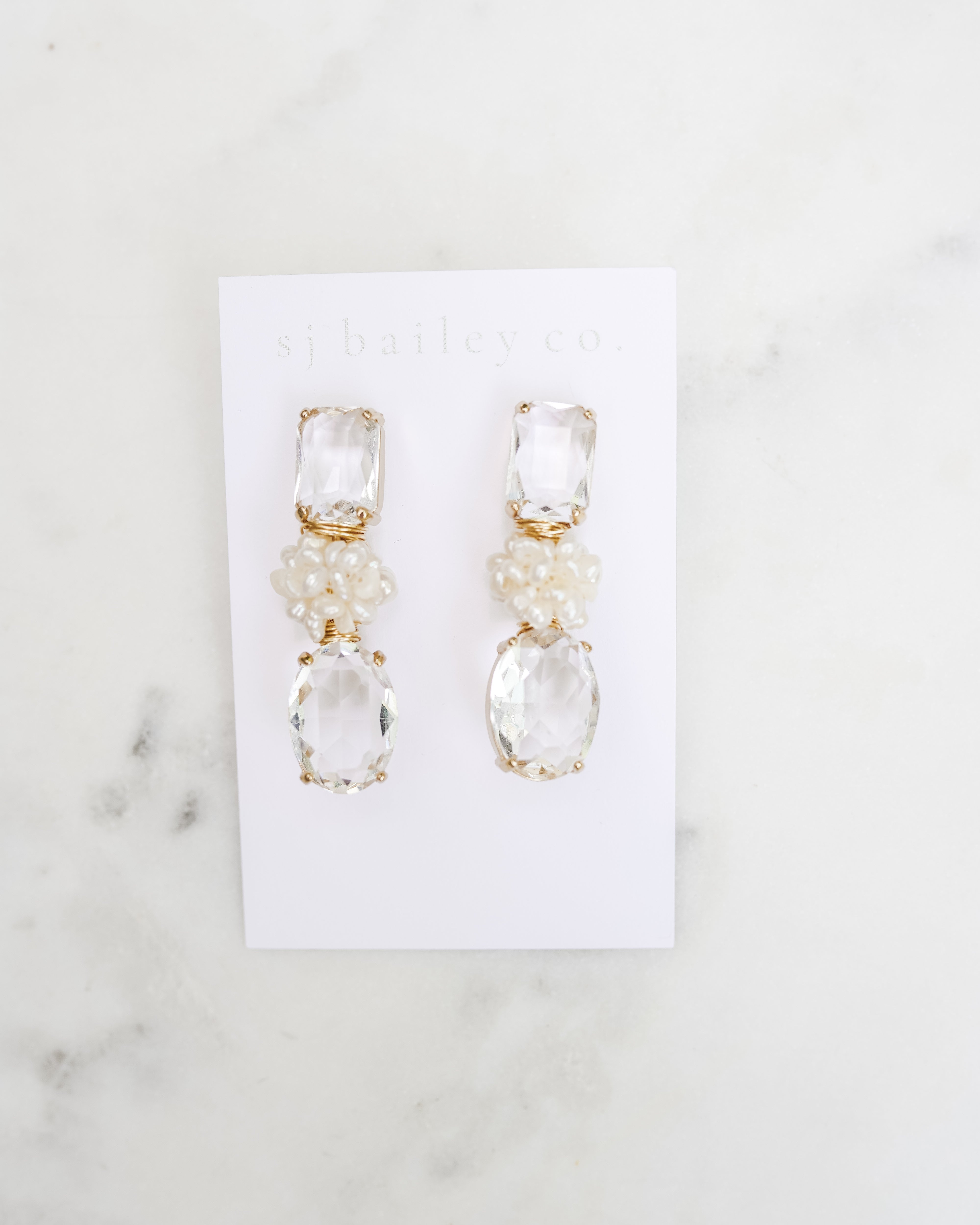 Crystal earrings with a pearl cluster. They are on a white card against a white marble background. 