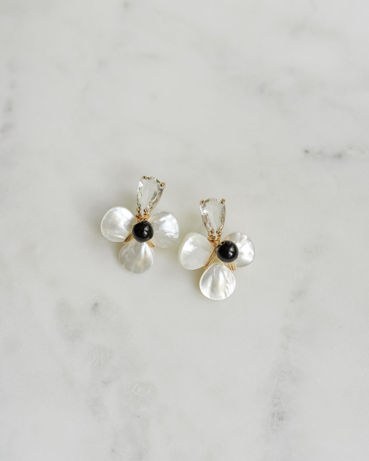 A pair of gold and pearl flower-shaped stud earrings with black bead centers, displayed on a light gray marble surface.