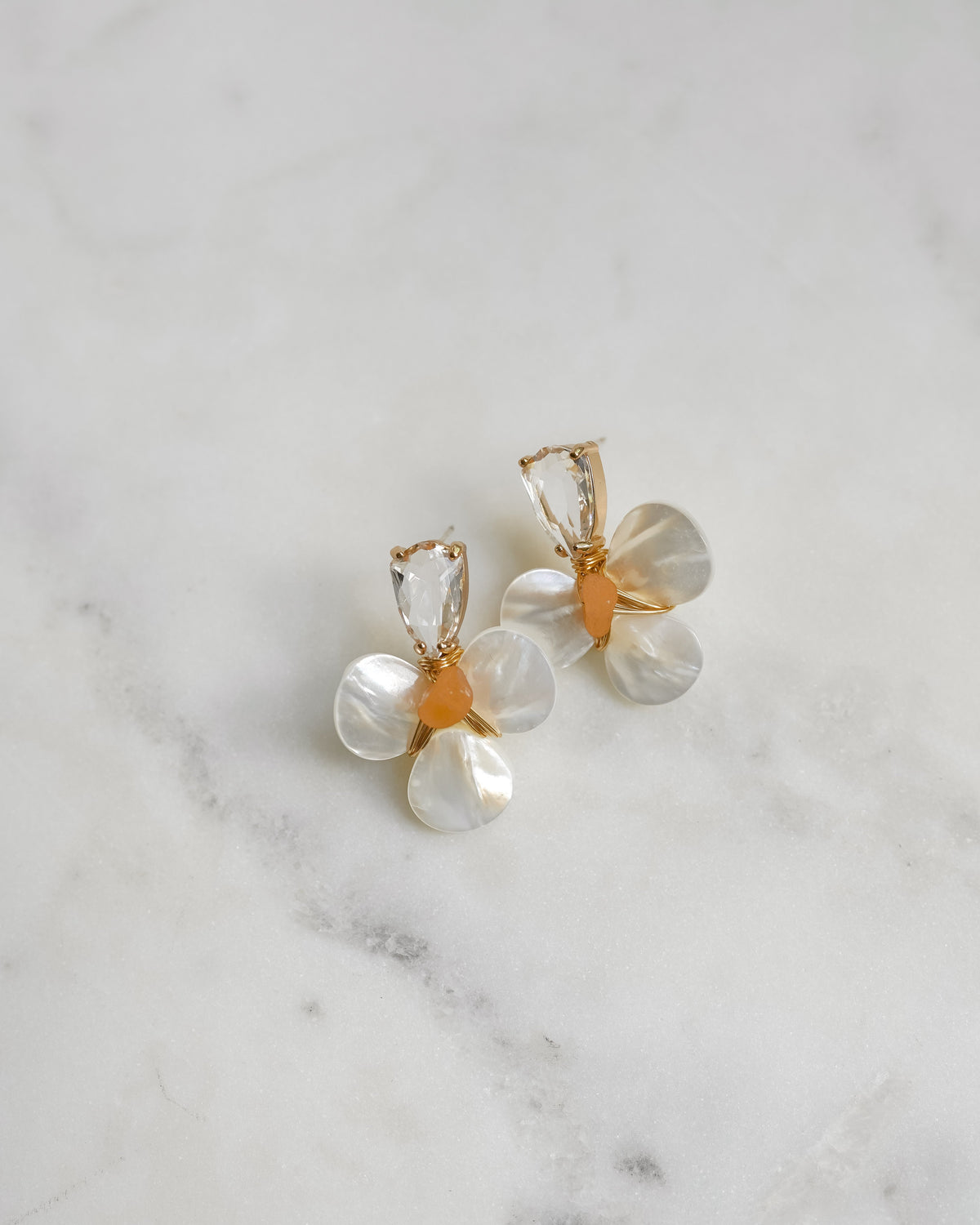 A pair of gold and white flower-shaped earrings with clear faceted stones, an orange beaded center and resting on a white marble surface.
