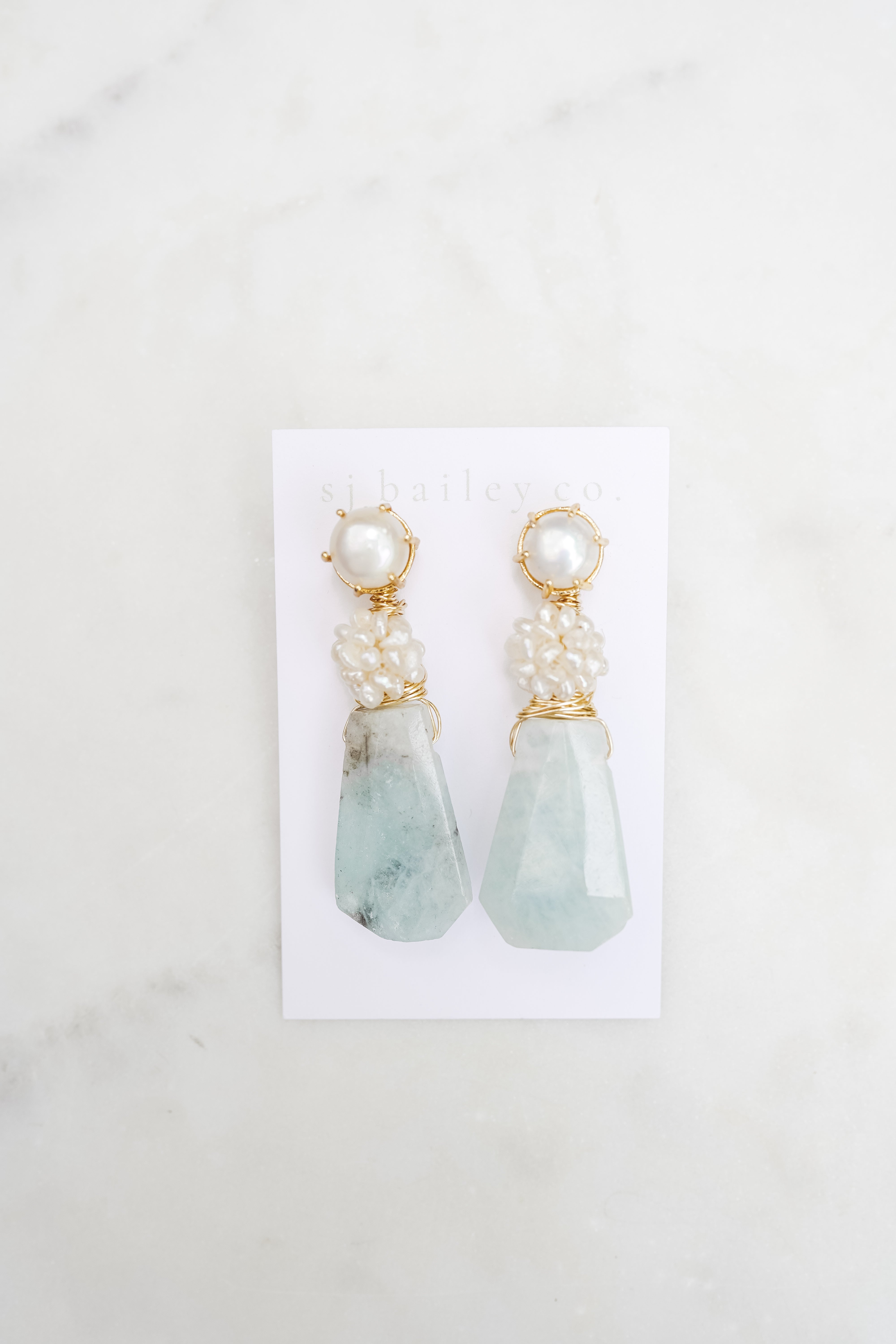 Pair of earrings with aquamarine teardrop stones and pearl-like tops on a white card against a light gray background.