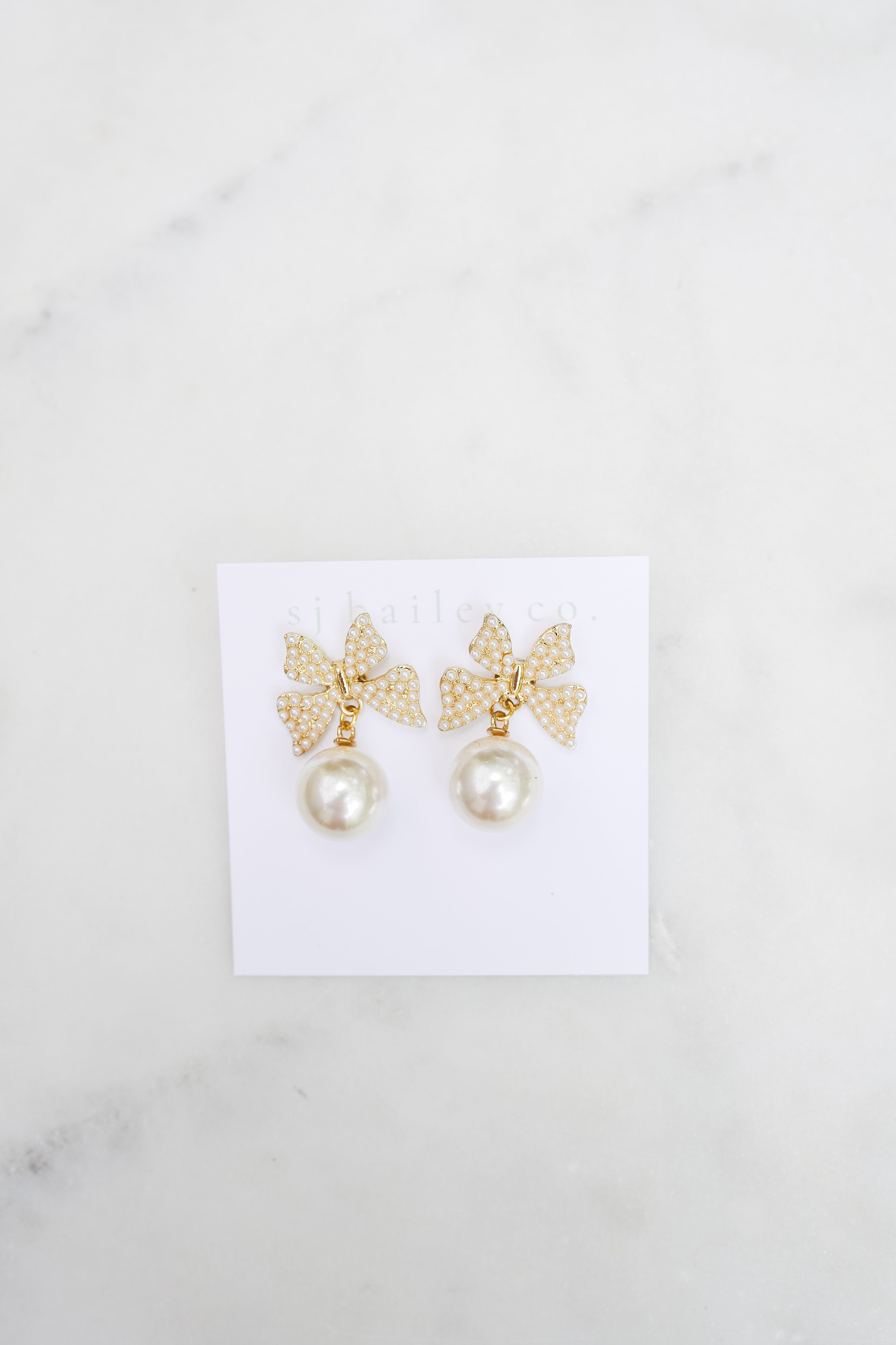 Golden and Pearl bow earrings with a pearl pendant on a white card. They are featured on a marble background. 