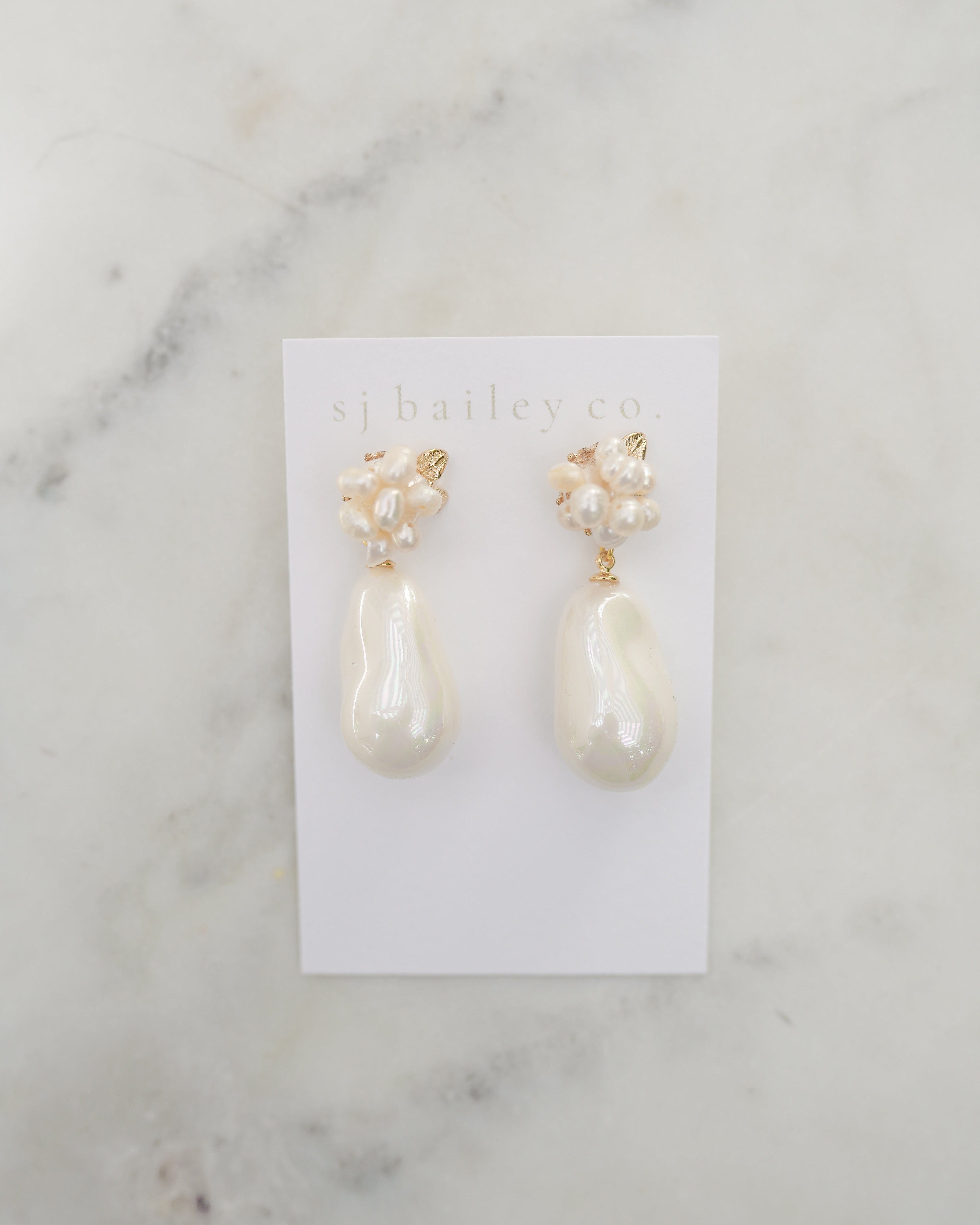 Pair of pearl earrings on a white card with 'sj bailey co' branding, against a marble background.