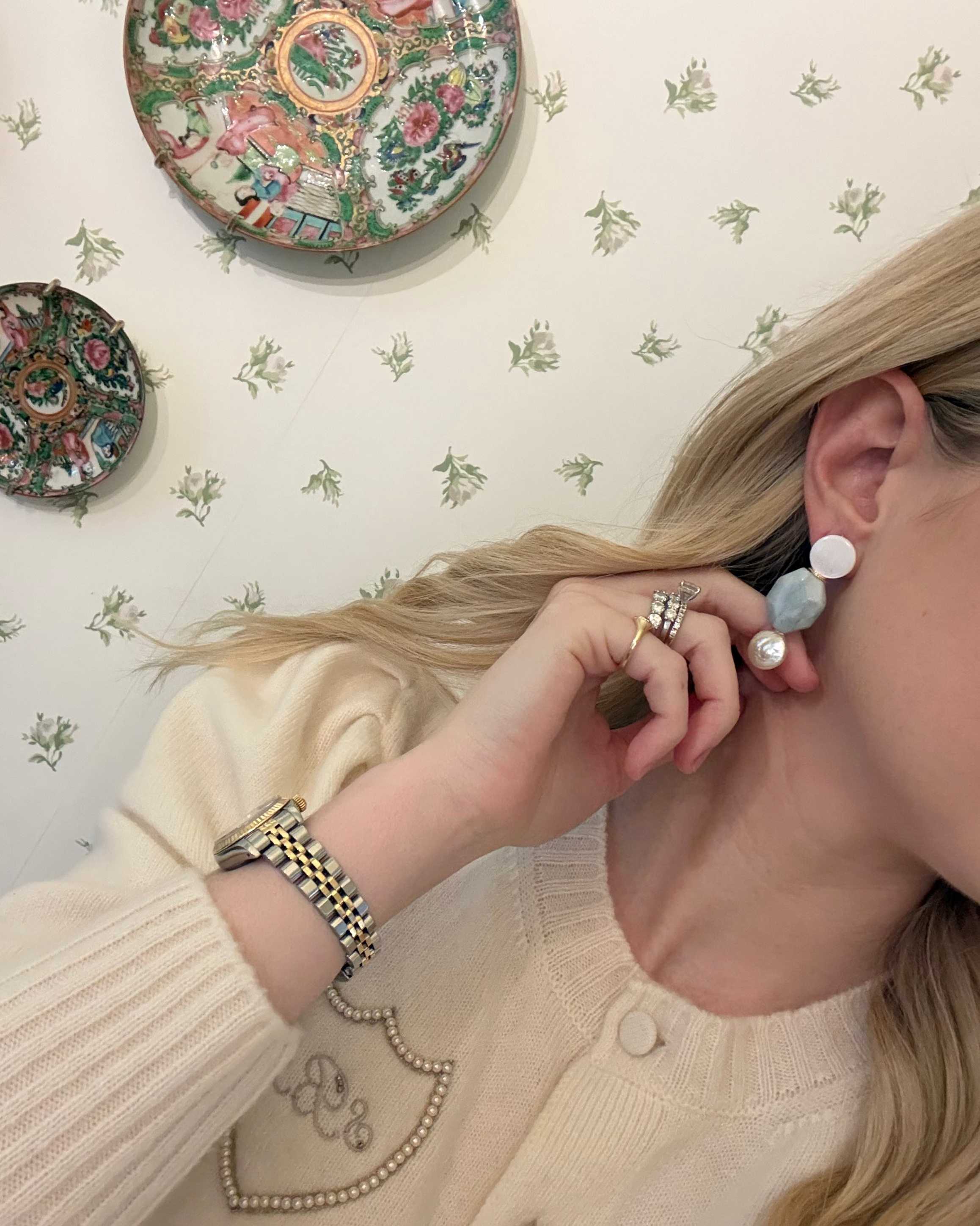 a blonde woman in a cream sweater wearing aquamarine and pearl statement earrings. she is in front of a floral wallpapered wall with green wedgwood and rose medallion plates on it