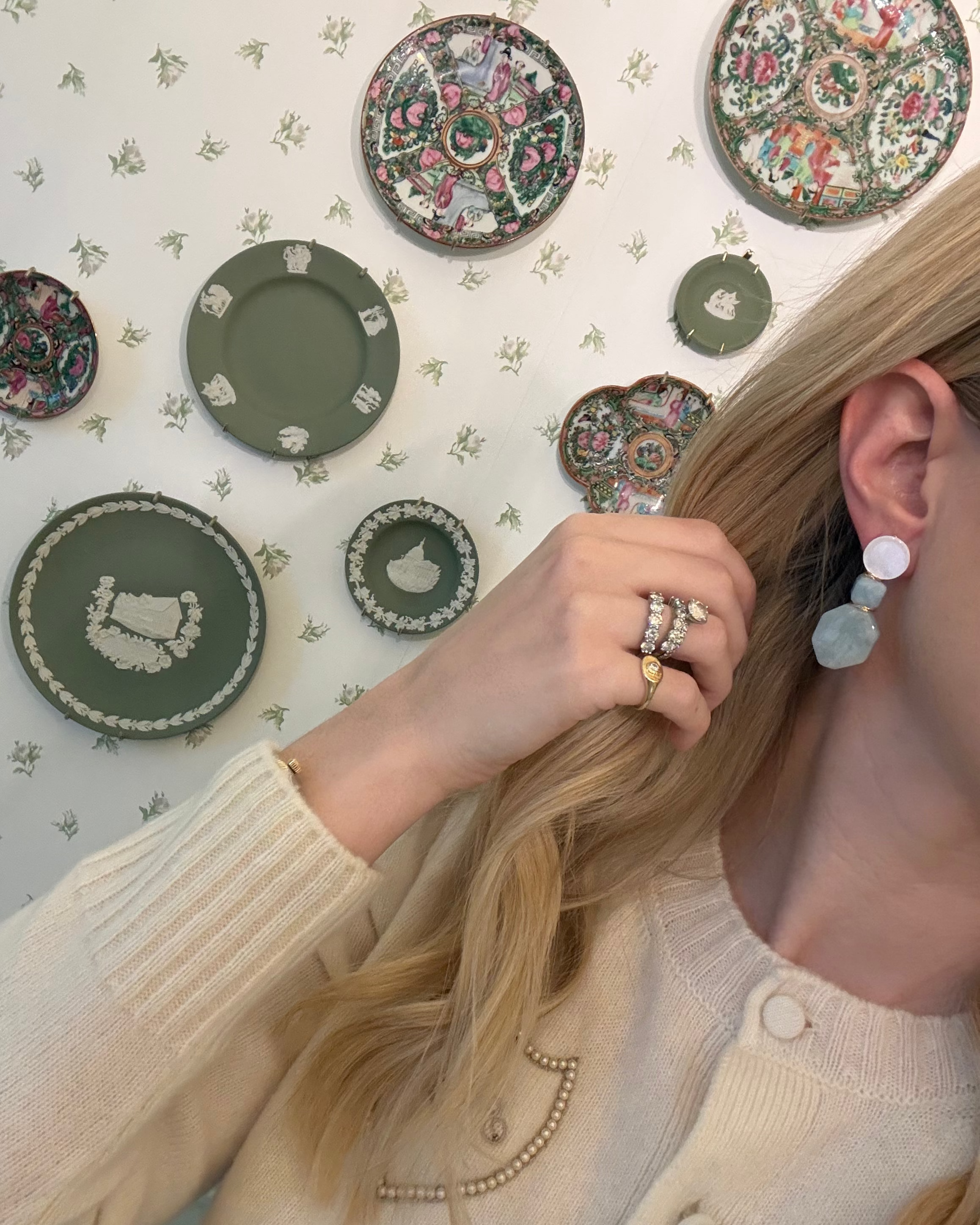 a blonde woman in a cream sweater wearing aquamarine and pearl statement earrings. she is in front of a floral wallpapered wall with green wedgwood and rose medallion plates on it