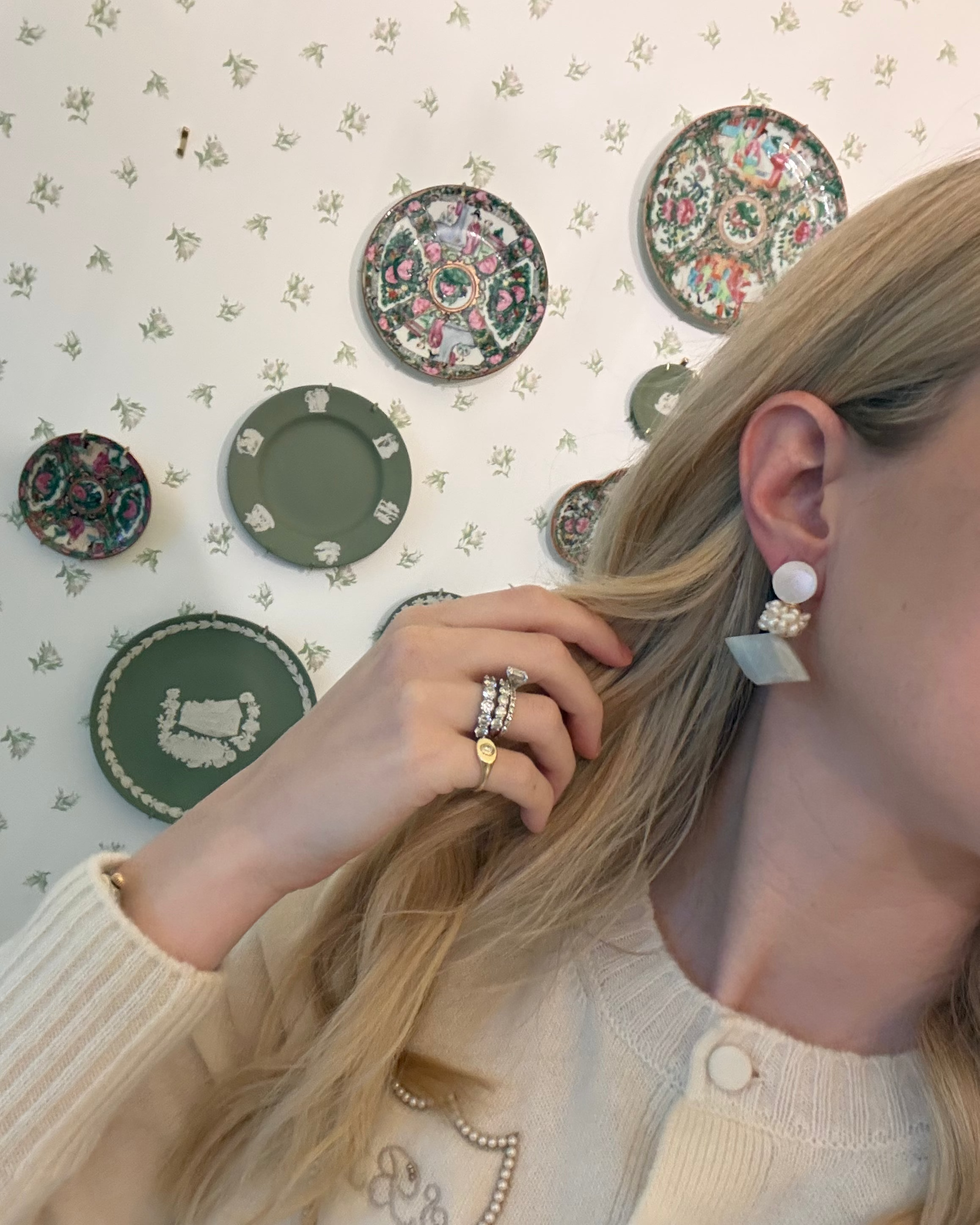 a blonde woman in a cream sweater wearing aquamarine and pearl statement earrings. she is in front of a floral wallpapered wall with green wedgwood and rose medallion plates on it