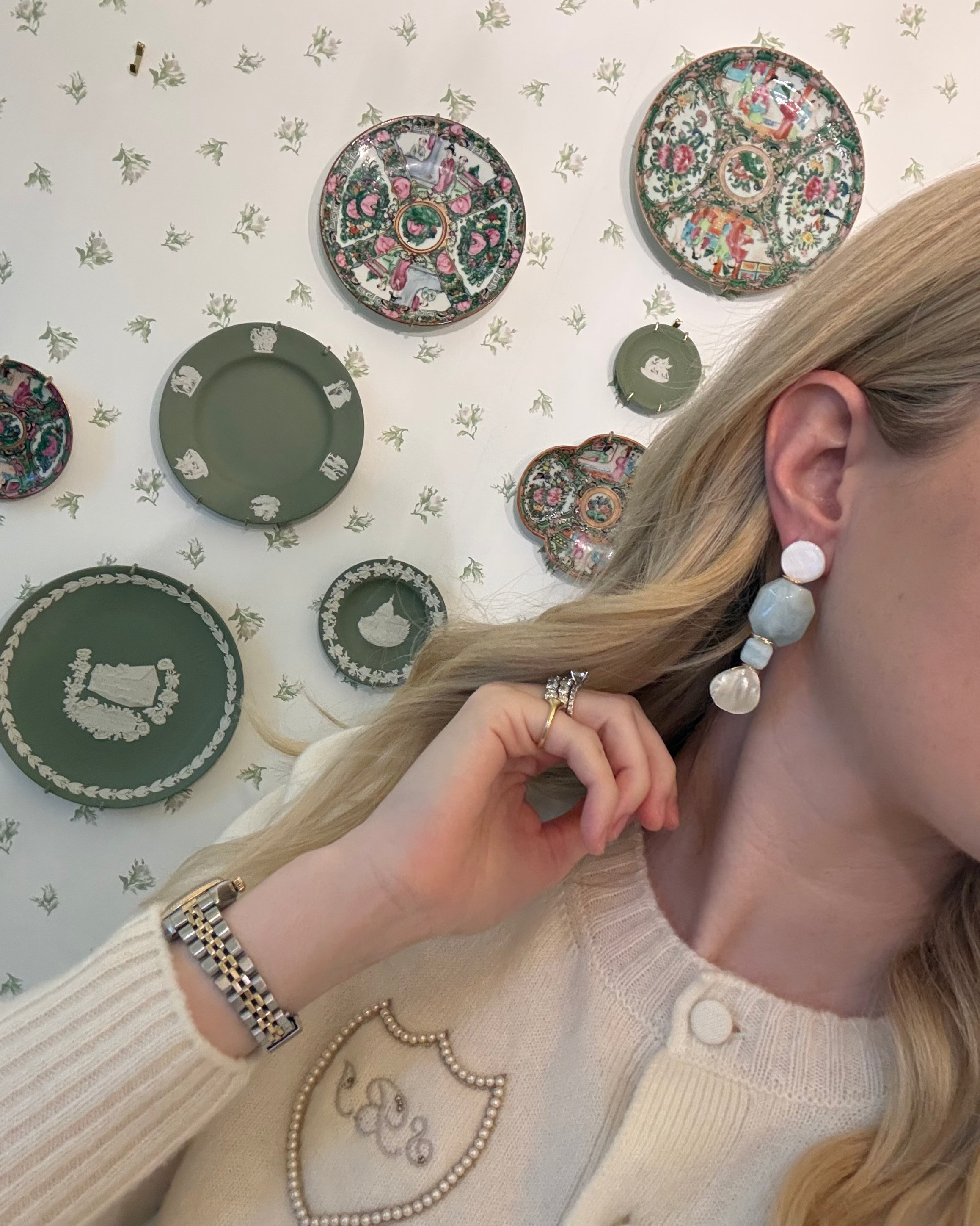 a blonde woman in a cream sweater wearing aquamarine and pearl statement earrings. she is in front of a floral wallpapered wall with green wedgwood and rose medallion plates on it