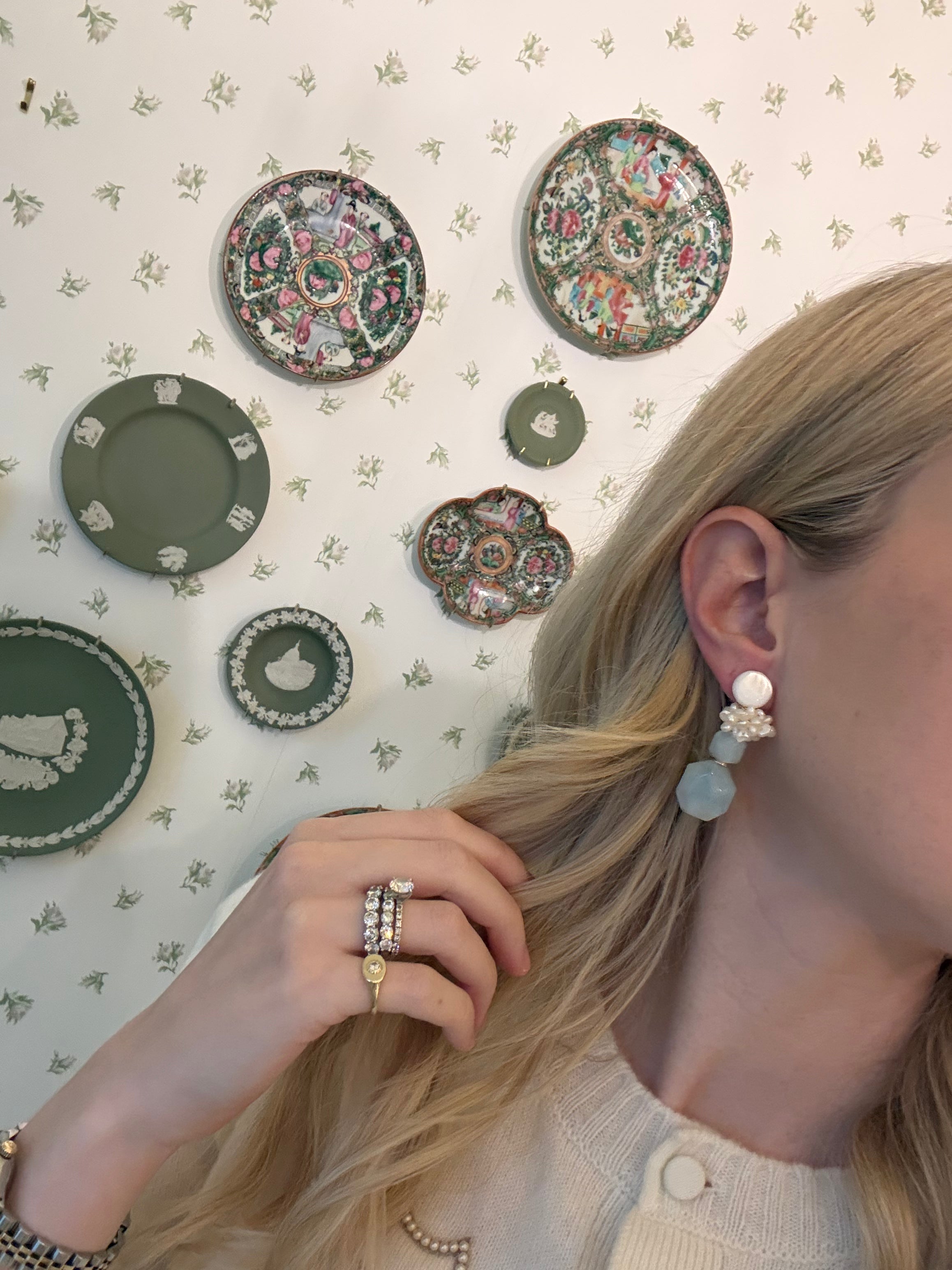 a blonde woman in a cream sweater wearing aquamarine and pearl statement earrings. she is in front of a floral wallpapered wall with green wedgwood and rose medallion plates on it
