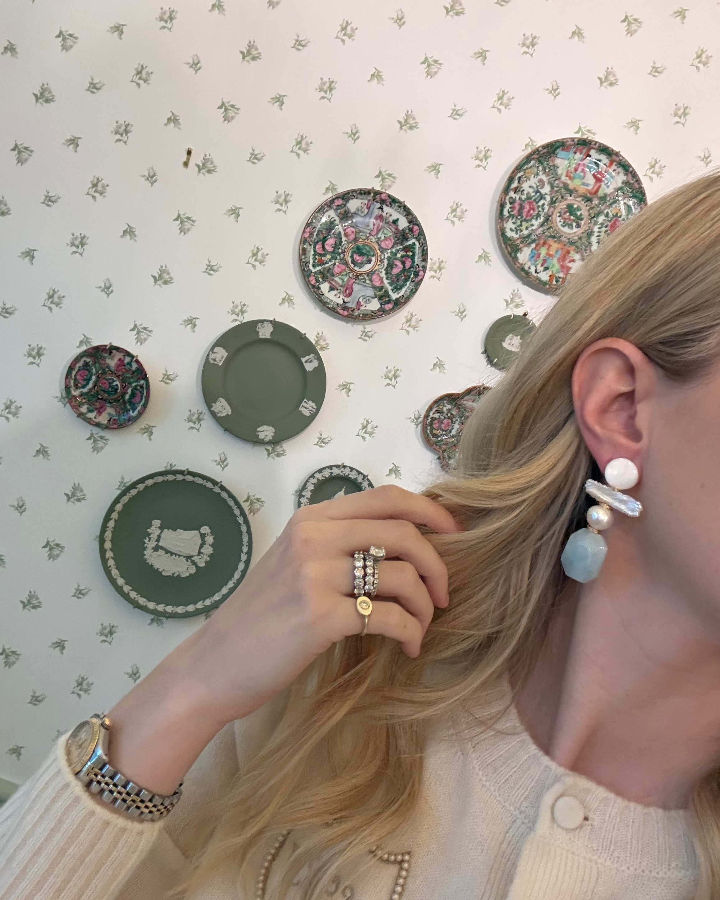 a blonde woman in a cream sweater wearing aquamarine and pearl statement earrings. she is in front of a floral wallpapered wall with green wedgwood and rose medallion plates on it